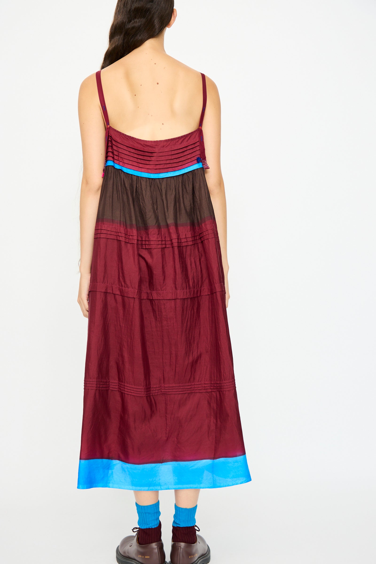 A person stands facing away, wearing the Injiri Silk and Cotton Slip Dress in Red—a relaxed fit, sleeveless dress with blue hem accents—paired with blue socks and brown shoes. Their long hair is worn to one side.