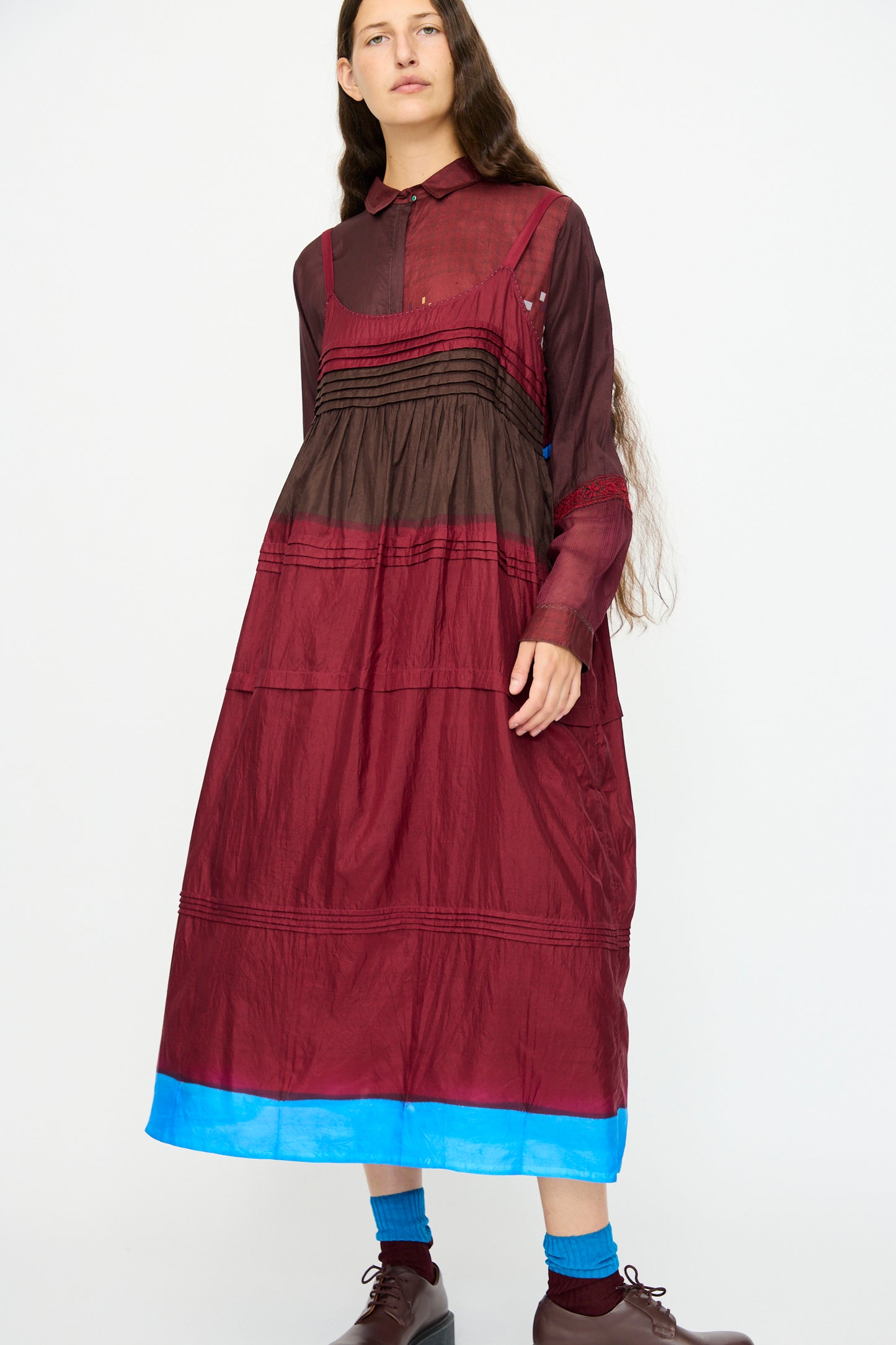 A person stands against a white background wearing the Injiri Silk and Cotton Slip Dress in Red, featuring a relaxed fit and multi-color details, paired with brown shoes and blue socks.