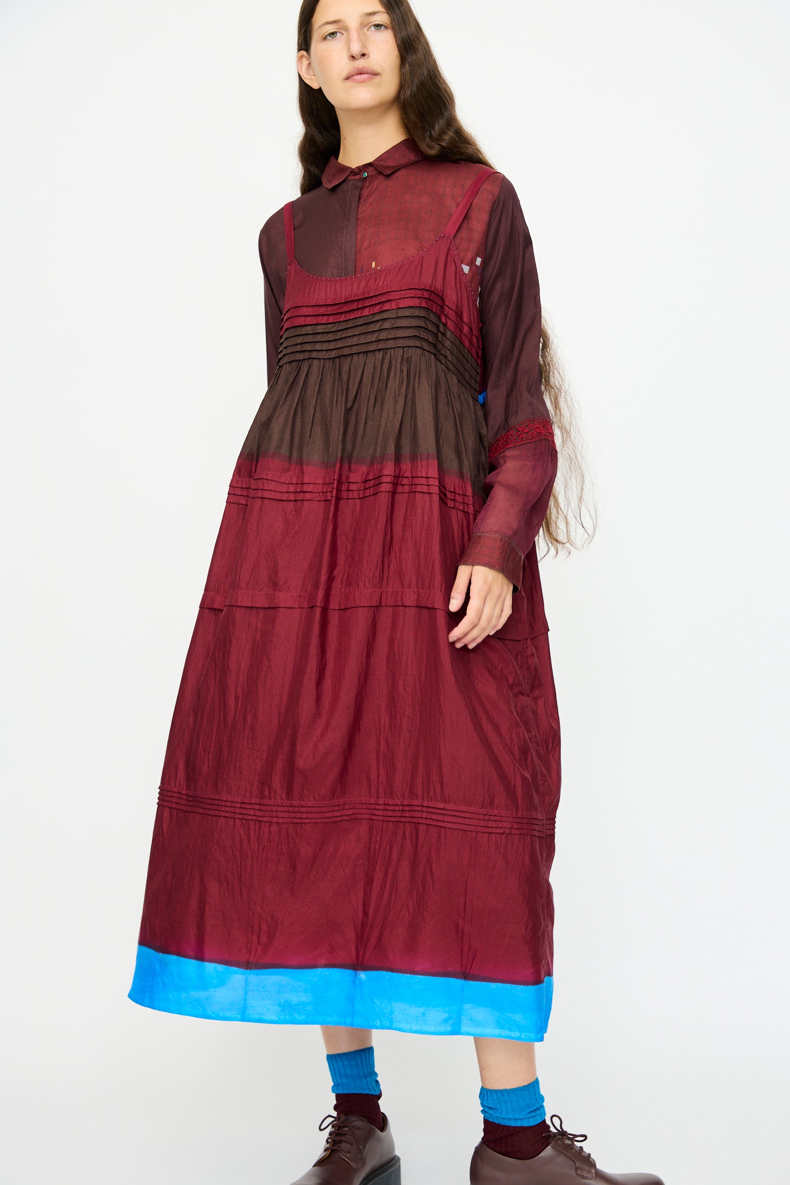 A person stands against a white background wearing the Injiri Silk and Cotton Slip Dress in Red, featuring a relaxed fit and multi-color details, paired with brown shoes and blue socks.