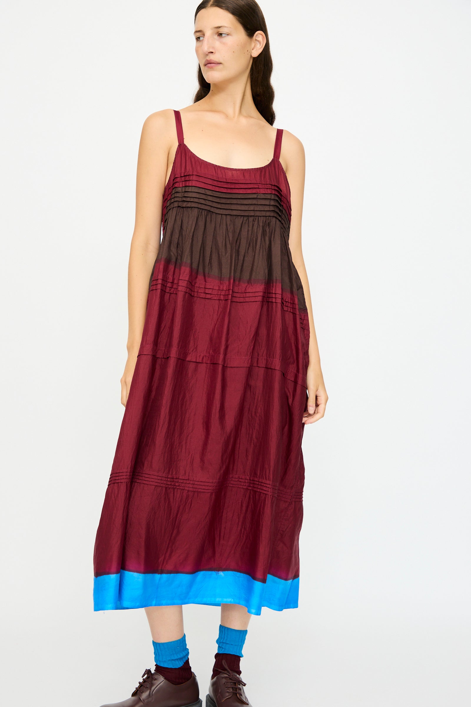 A woman wears the Injiri Silk and Cotton Slip Dress in Red, featuring a relaxed fit with burgundy, black, and bright blue accents, styled with brown shoes and blue socks.