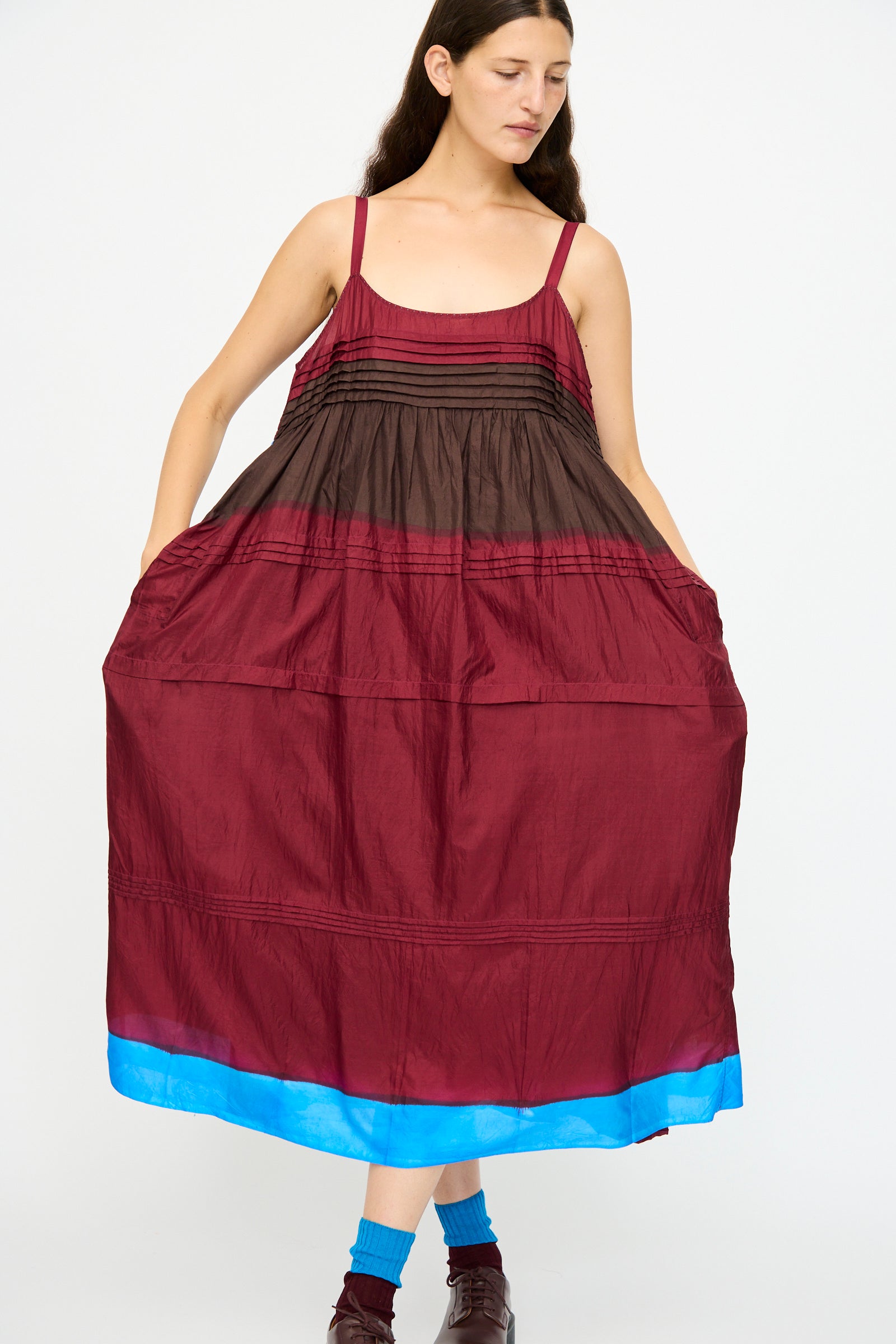 A woman models the Injiri Silk and Cotton Slip Dress in Red, a relaxed-fit, multi-color piece with brown and burgundy sections and a blue hem, styled with blue socks and brown shoes.
