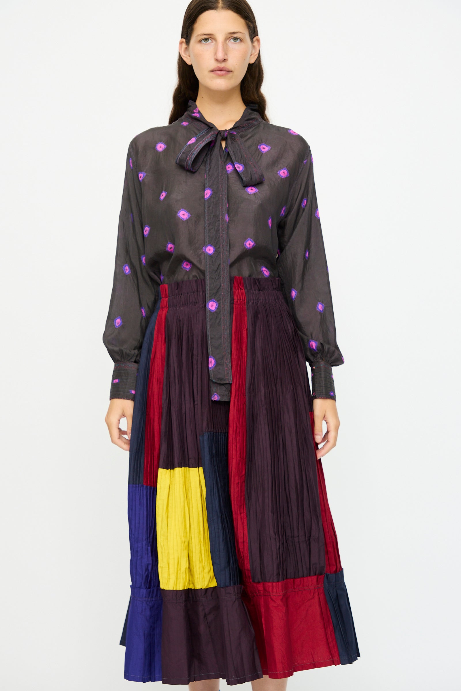 A woman stands against a white background wearing a sheer black blouse with purple floral patterns and a bow at the neck, paired with the Injiri Taffeta Silk Skirt in Multi.