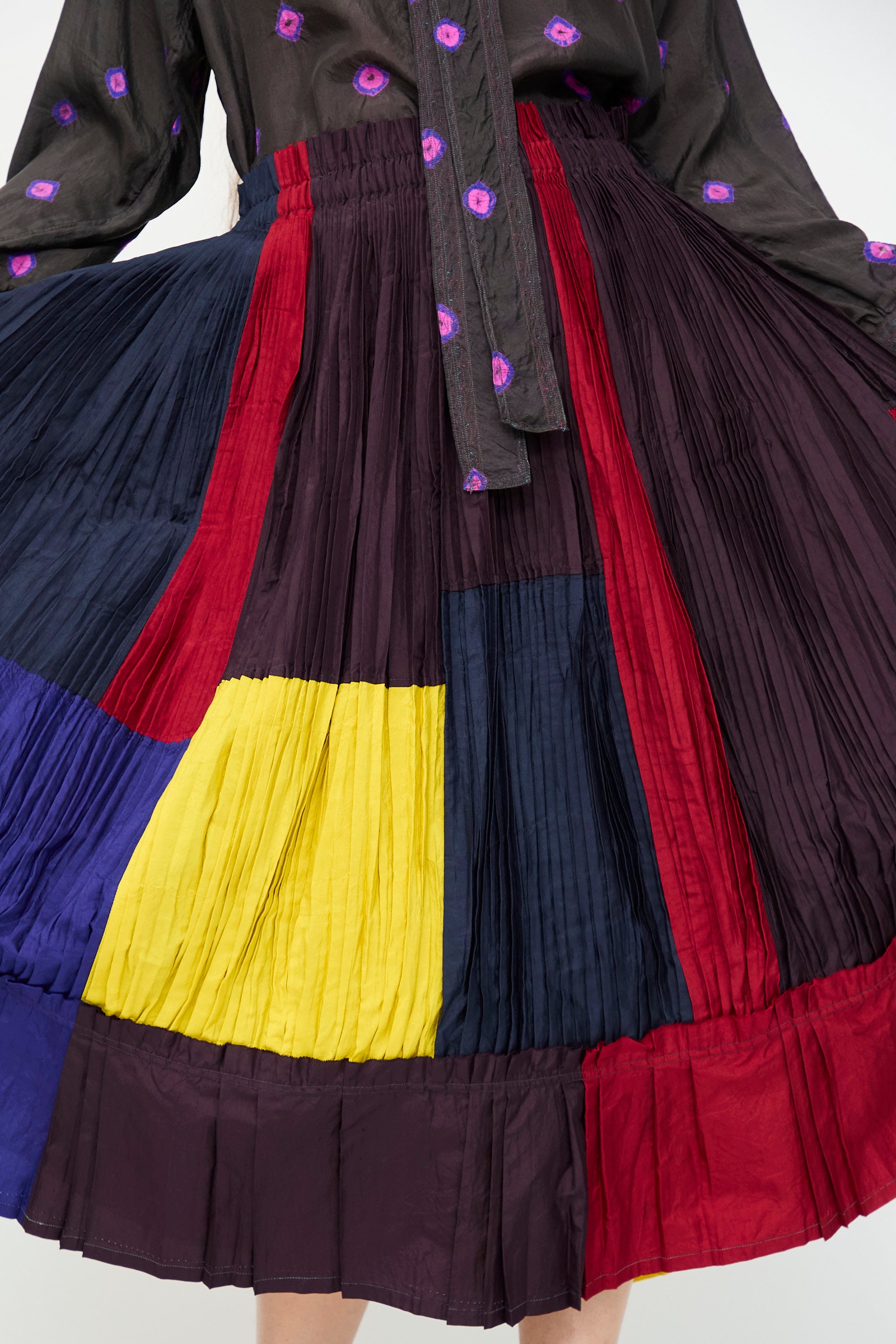 A person wearing the Injiri Taffeta Silk Skirt in Multi, a color-block pleated midi with bold red, yellow, navy, black, and purple panels, paired with a black top featuring purple floral accents.