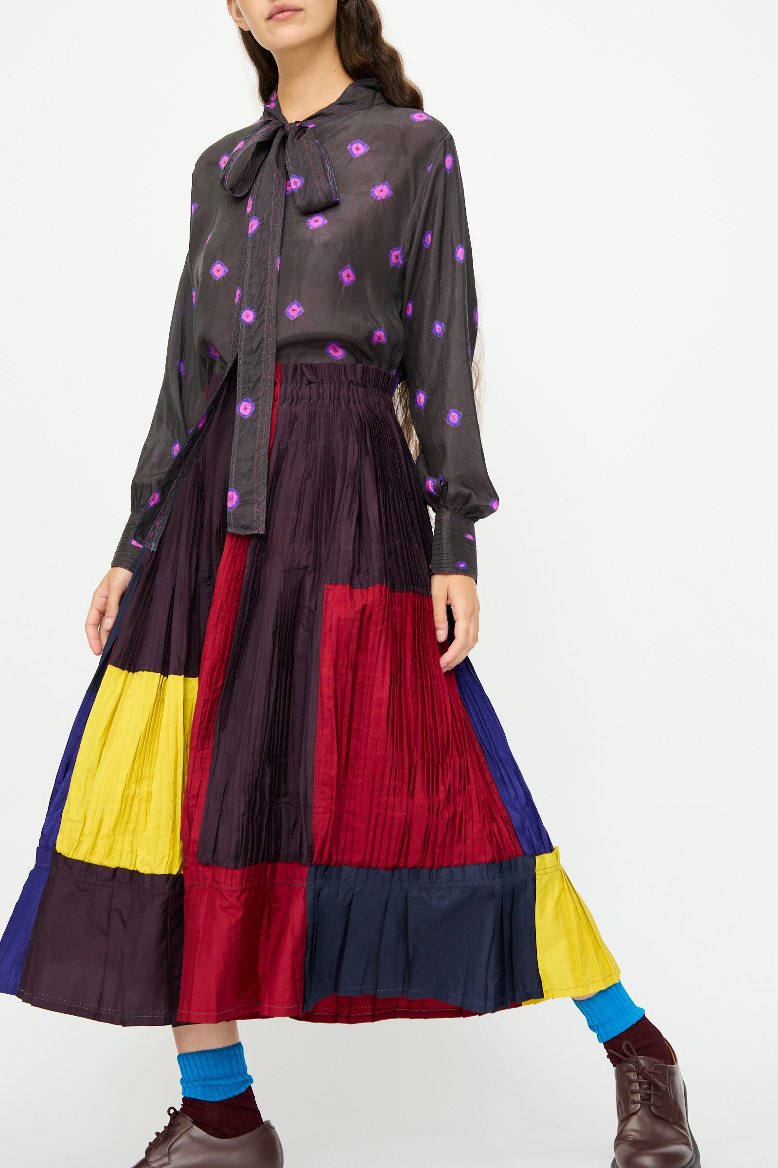 A woman wears a black blouse with purple dots, a bow at the neck, and Injiri’s Taffeta Silk Skirt in Multi, finished with brown shoes and blue socks.
