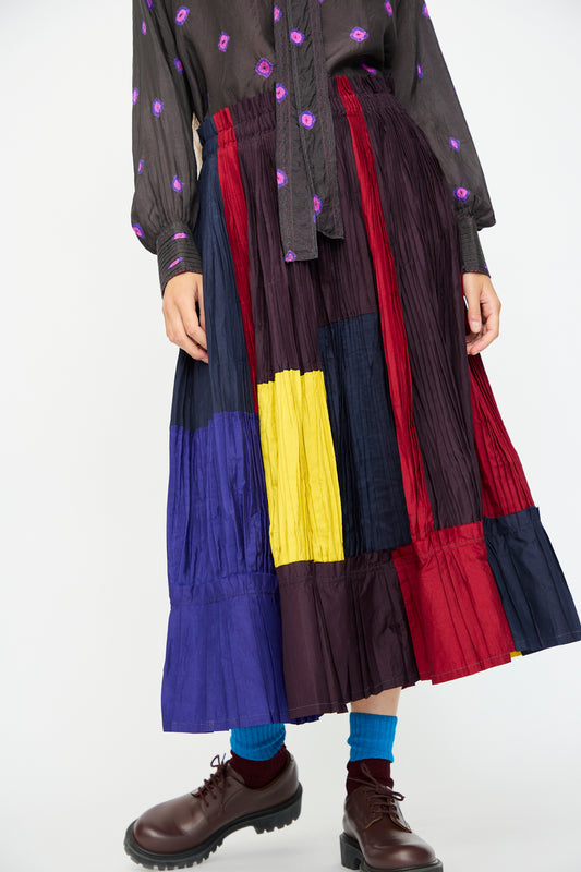 A person wears the Injiri Taffeta Silk Skirt in Multi, featuring red, yellow, blue, and black panels in an A-line silhouette, styled with a dark shirt with purple patterns, blue socks, and brown shoes.
