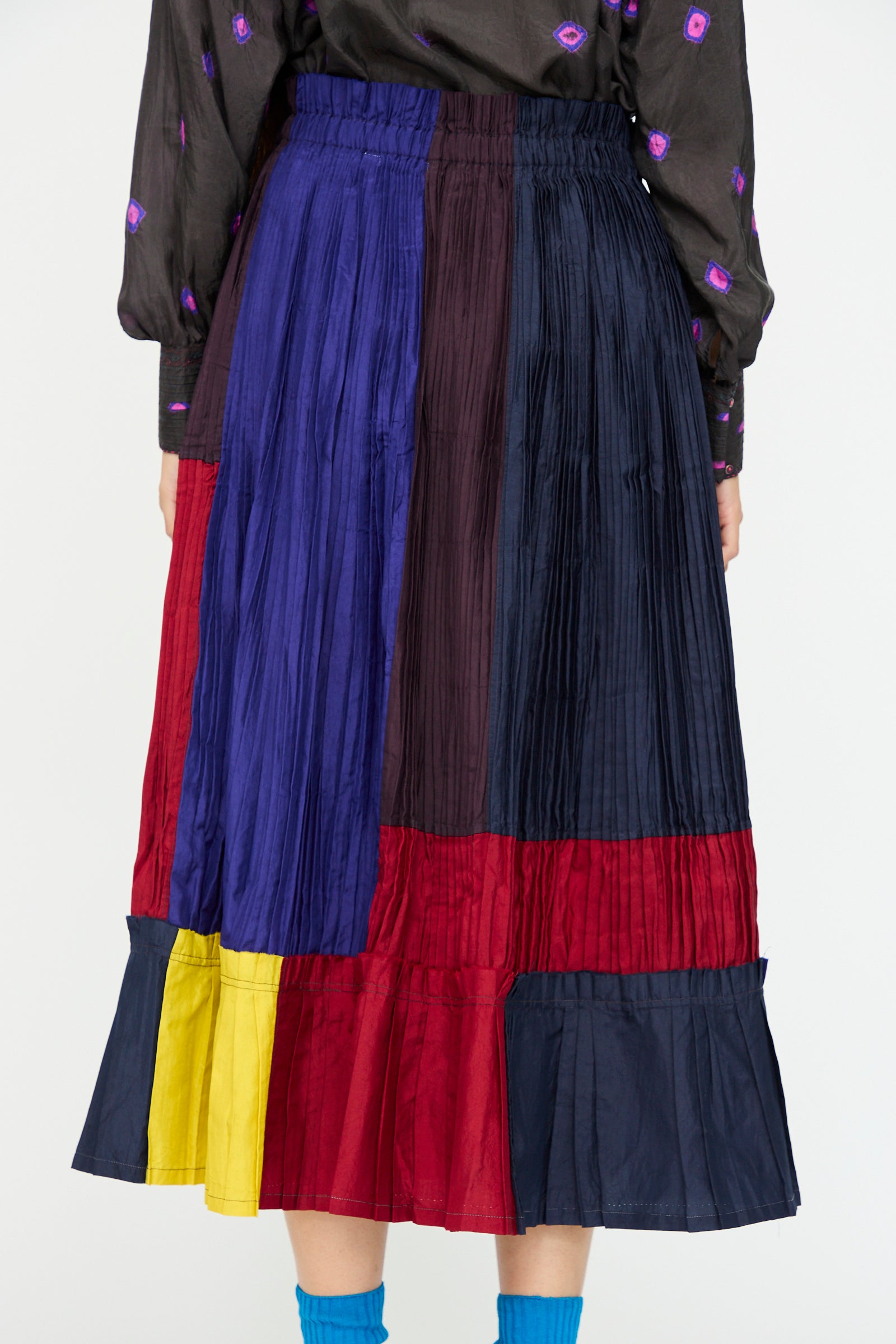 A person wearing the Injiri Taffeta Silk Skirt in Multi—a vibrant, pleated color-block midi—paired with a dark patterned top and blue socks.