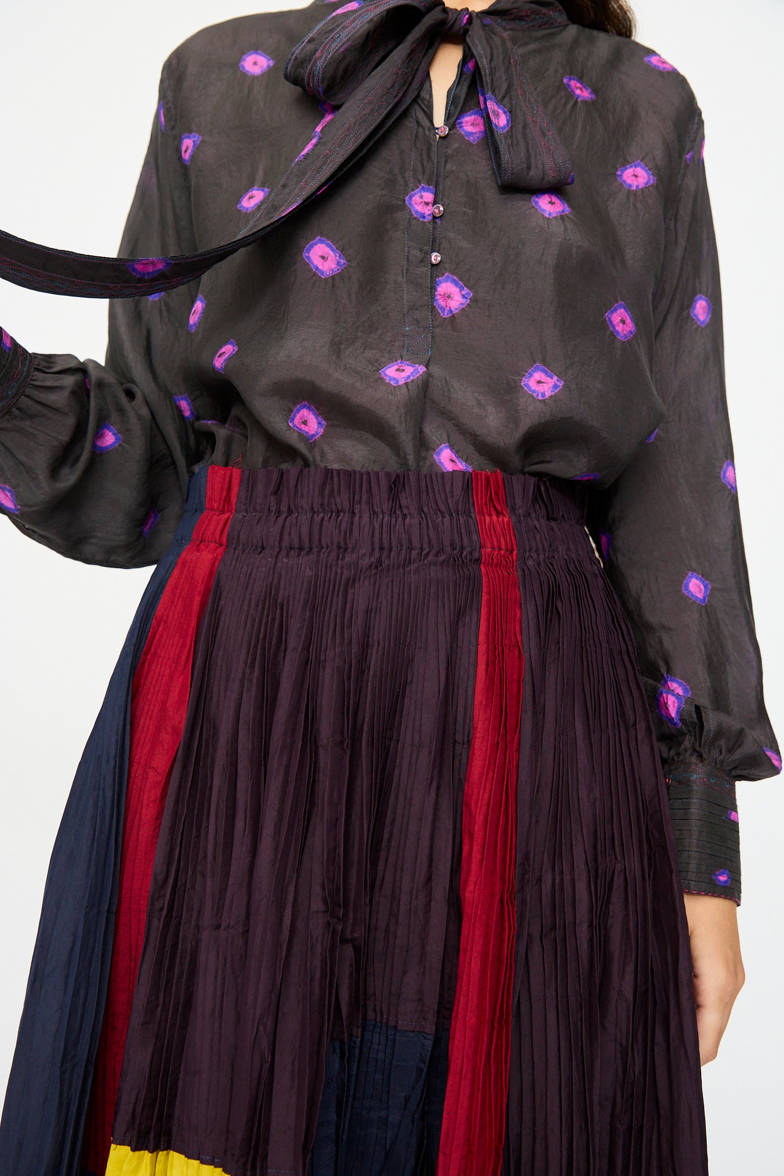 A person wearing a black blouse with purple spots and a large neck bow, paired with the Injiri Taffeta Silk Skirt in Multi, which features color-blocked pleated panels of burgundy, red, navy, and yellow.