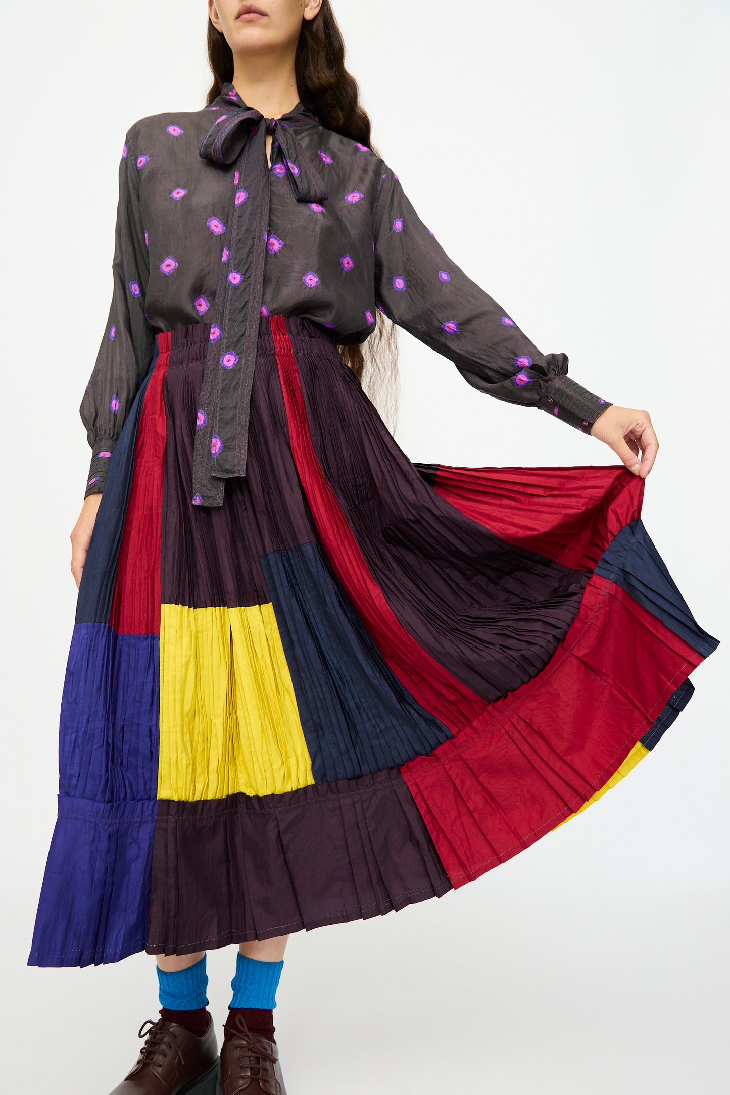 A woman wears a dark blouse with a bow and purple floral print, paired with the Injiri Taffeta Silk Skirt in Multi—a color-block A-line skirt in red, yellow, blue, and maroon tones—and brown shoes.