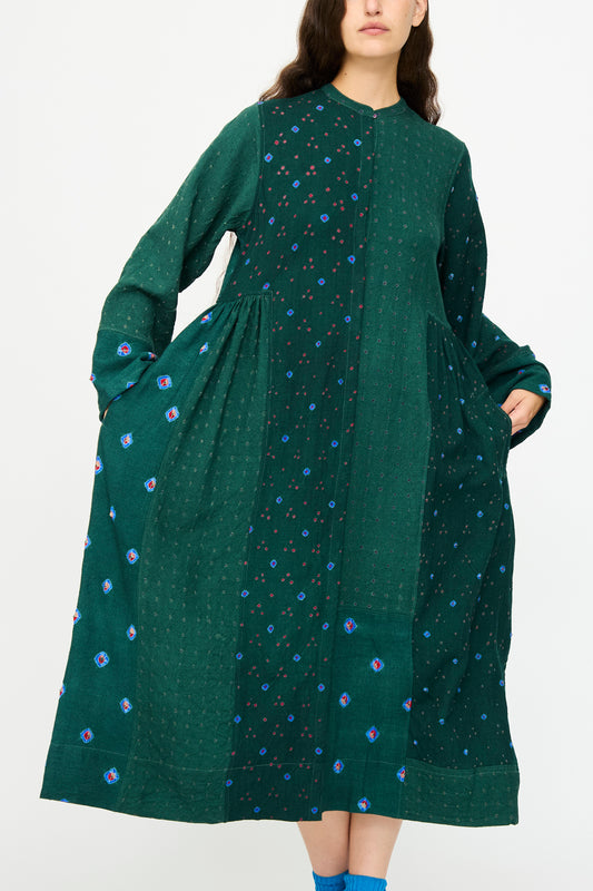 A woman wearing the Injiri Wool Dress in Green, a long-sleeve patchwork style featuring blue and pink small floral patterns, stands with her hands in the side pockets.
