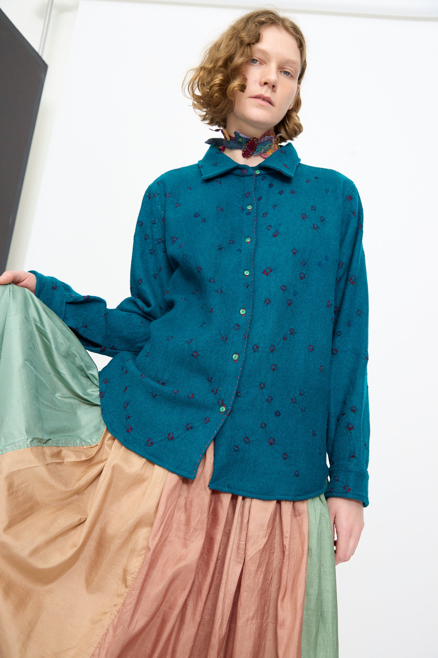 A person stands indoors against a light background, wearing a teal button-up shirt with embroidered details and a multicolored, flowing skirt, accessorized with the Injiri Wool Silk Neckband in Multi.