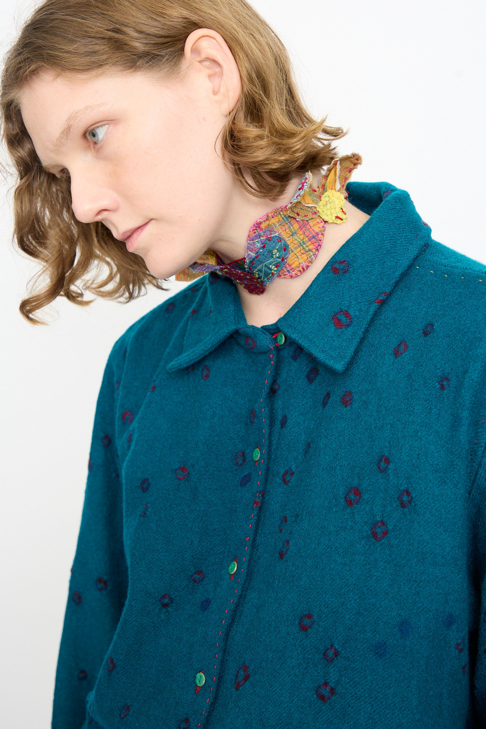 A person with light brown hair is wearing a teal button-up shirt featuring a patterned design and accessorizing with a colorful, crocheted fish choker necklace layered with the unique Wool Silk Neckband in Multi by Injiri.
