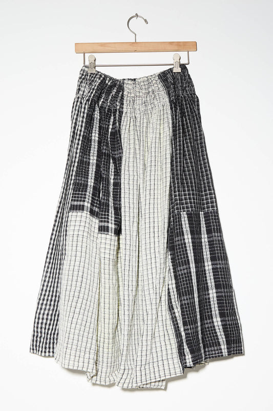 The Wool and Silk Skirt in Check by Injiri features a full skirt with an elastic waistband, showcasing a patchwork design of black and white checkered and striped patterns. It's displayed on a wooden hanger against a white background.