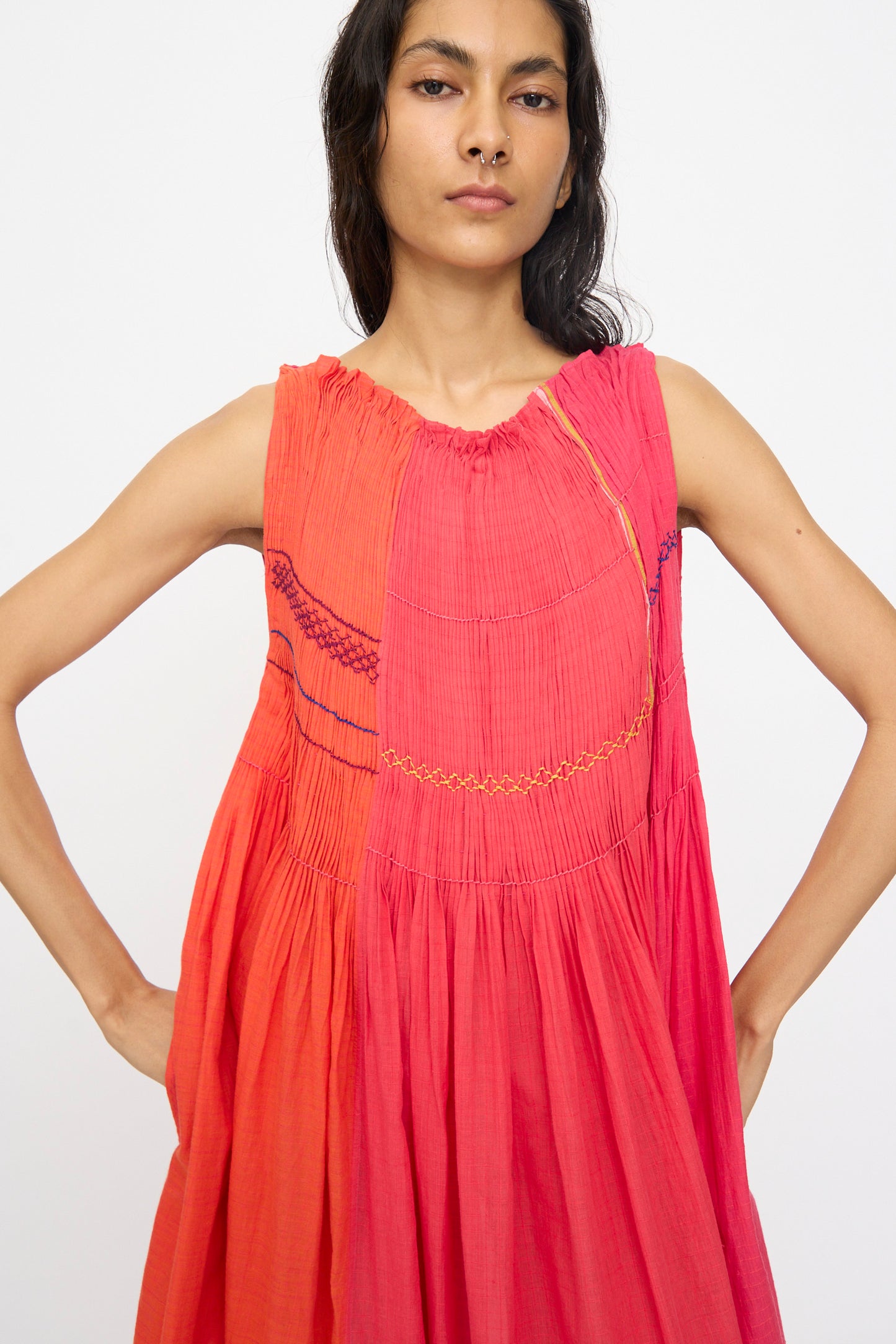 A woman wears the Injiri Woven Cotton Dress in Pink and Red, featuring embroidered details, while standing against a plain white background.