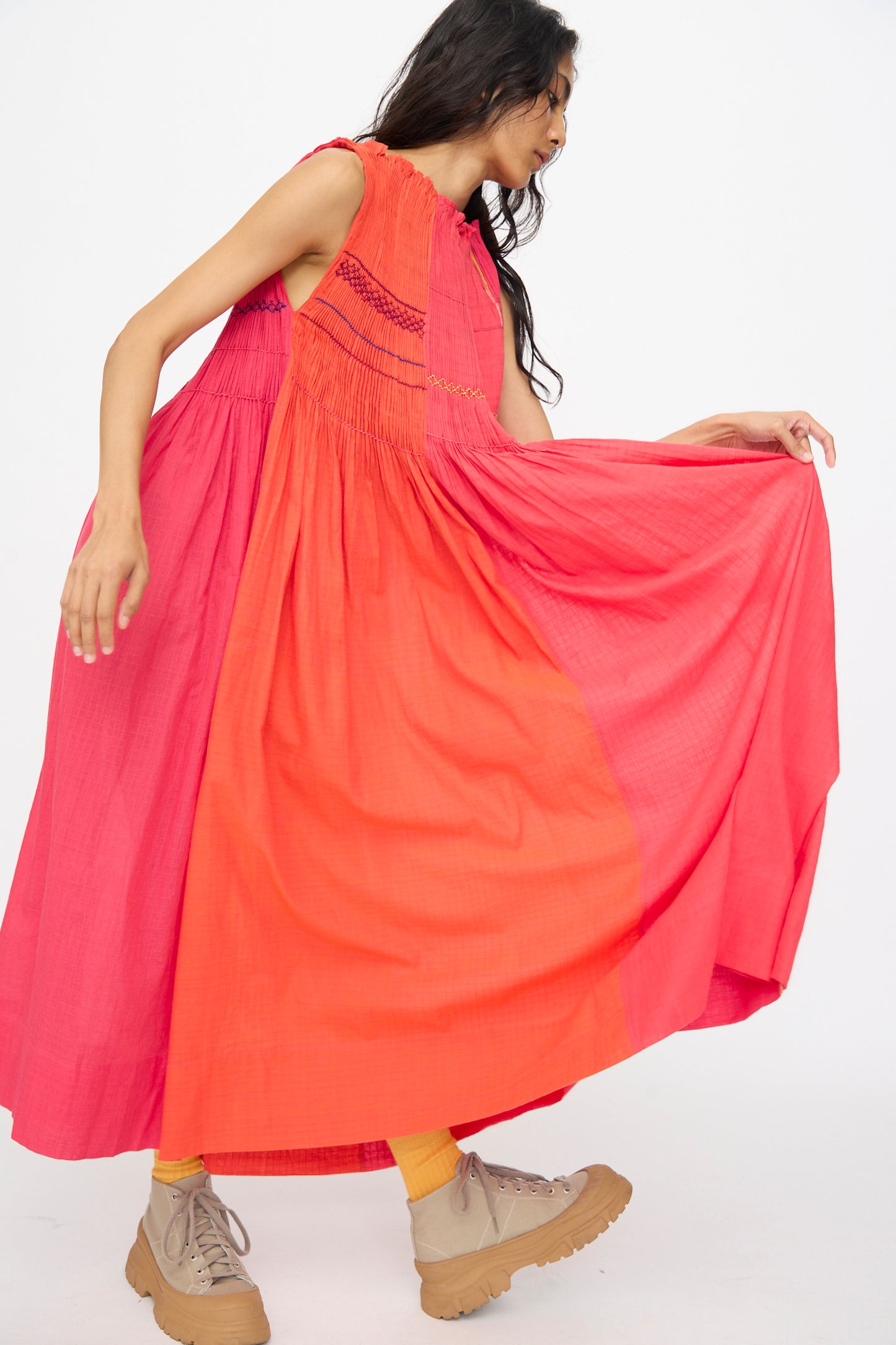 A woman wears the Injiri Woven Cotton Dress in Pink and Red, a loose, sleeveless midi style. She lifts one side of the skirt, paired with tan platform shoes and yellow socks against a plain white background.
