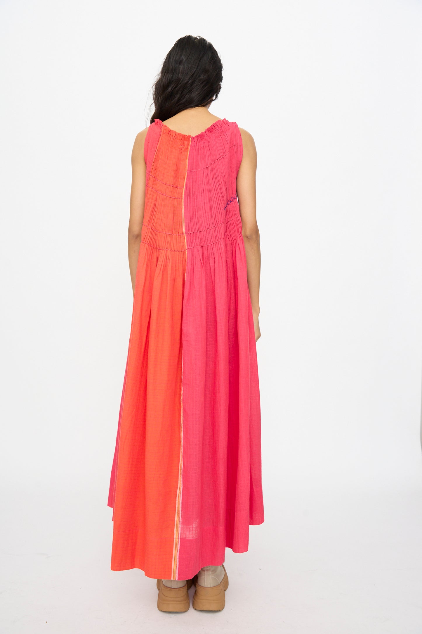 A person with long dark hair stands facing away, wearing the Injiri Woven Cotton Dress in Pink and Red, which features a sleeveless design and gathered waist, paired with tan platform shoes against a plain white background.