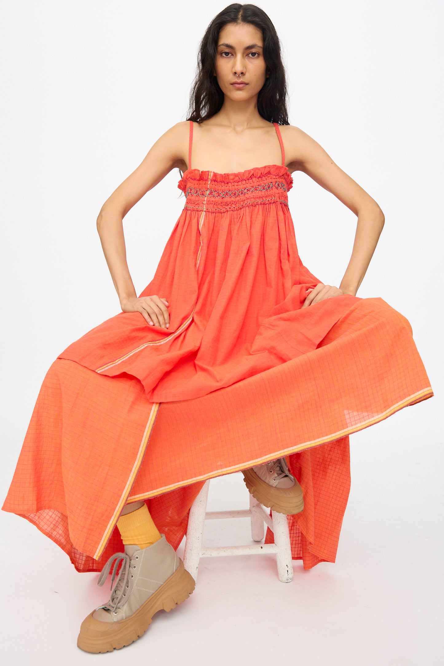Someone sits on a white stool against a plain white background, wearing the Injiri Woven Cotton Sleeveless Top in Red styled as a loose dress, paired with beige lace-up boots and yellow socks.