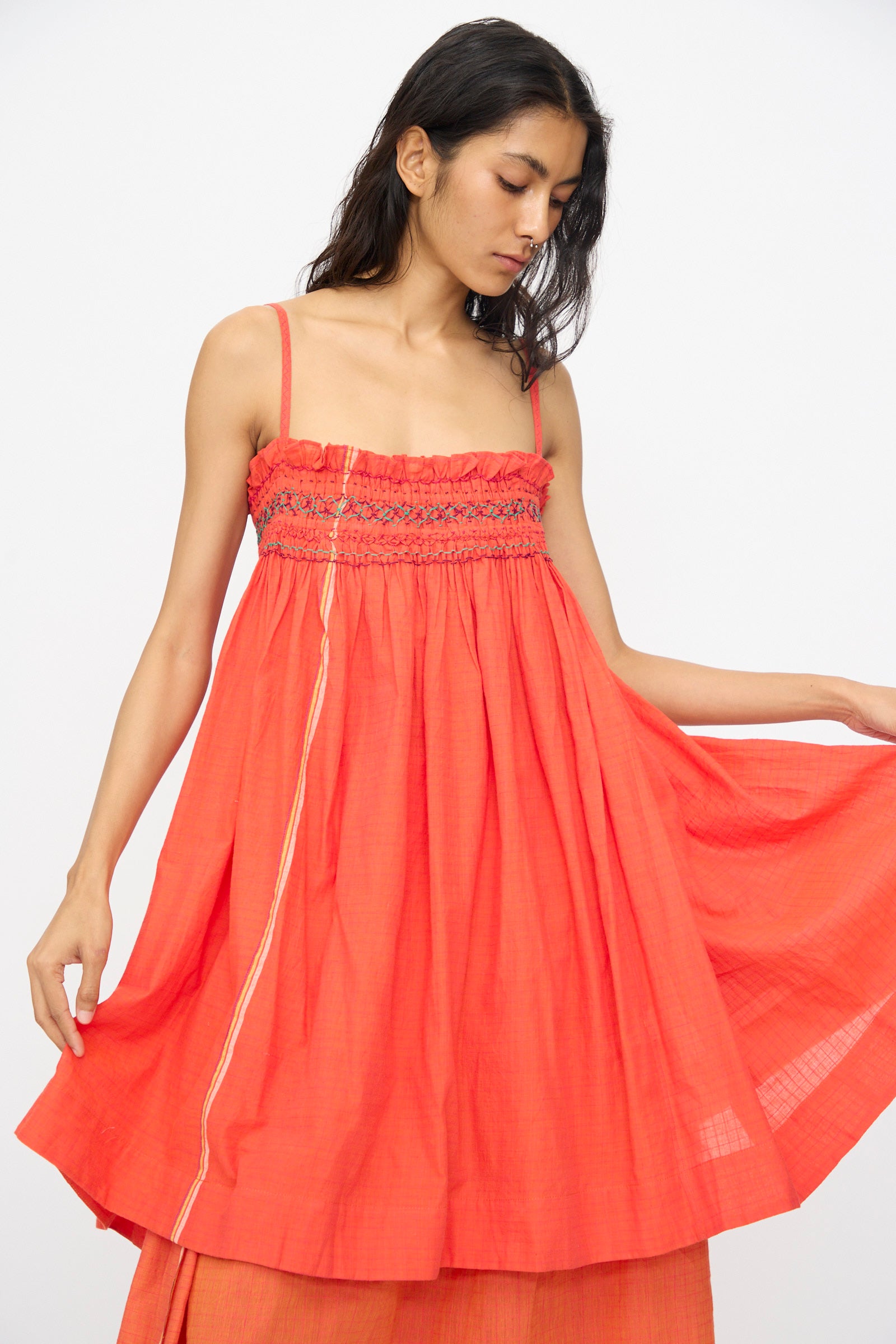 A woman wears the Injiri Woven Cotton Sleeveless Top in Red, featuring a loose fit, smocked details, and an embroidered square neck, holding out the skirt of her dress with one hand.