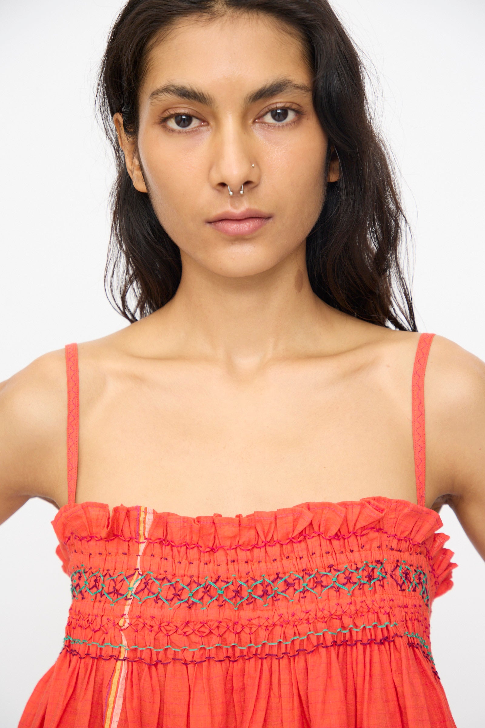 A woman with long dark hair faces the camera against a plain background, wearing the Injiri Woven Cotton Sleeveless Top in Red, styled as a smocked dress with thin straps and an embroidered square neckline.