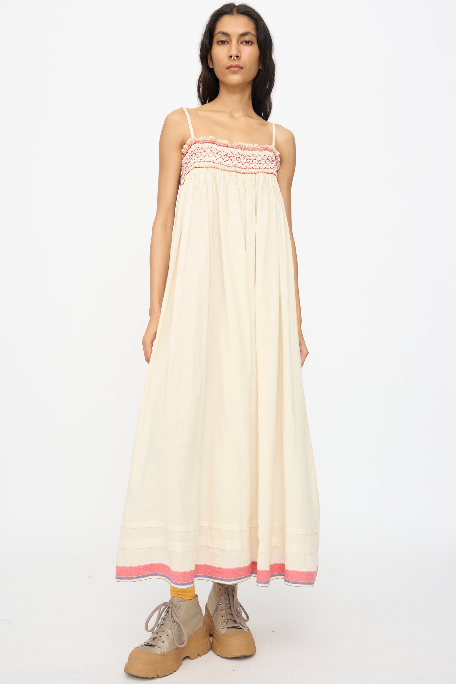 A person stands facing forward in the Injiri Woven Cotton Slip Dress in White with Red Stitch, paired with tan boots and yellow socks, against a plain white background.