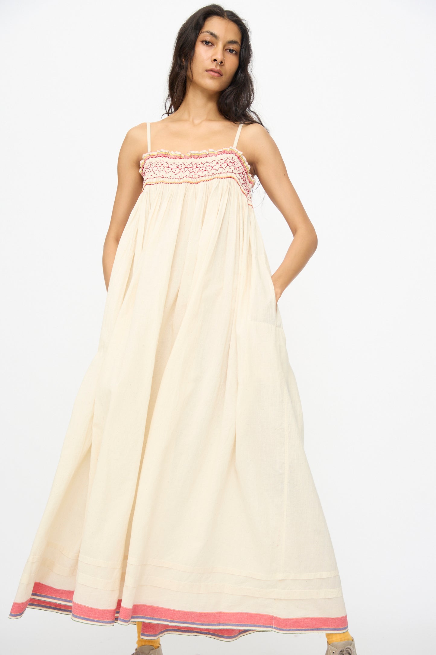 A woman wears the Injiri Woven Cotton Slip Dress in White with Red Stitch, a long, sleeveless piece with red detailing and pink trim, standing against a white background.