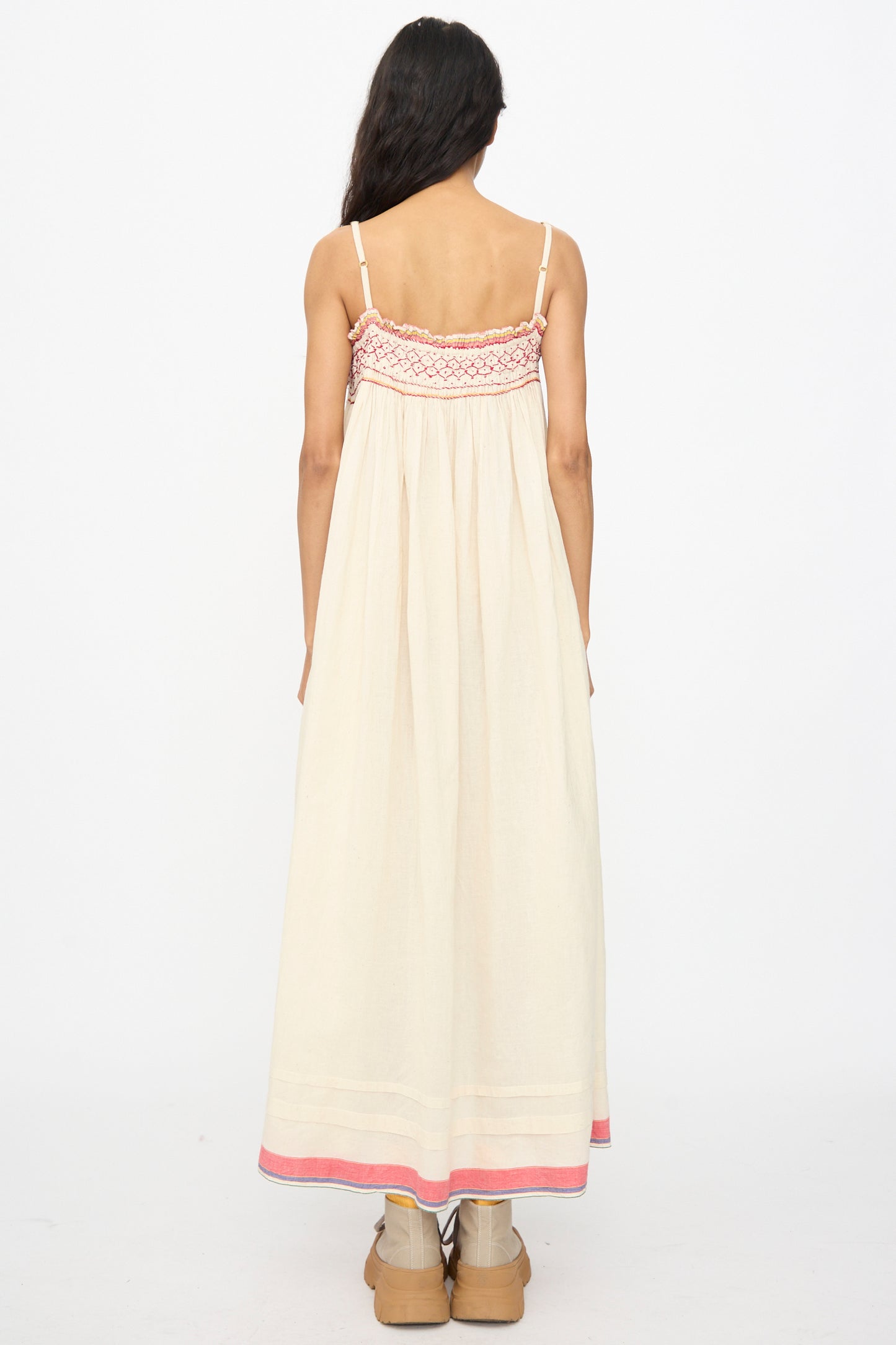 A woman stands with her back to the camera, wearing the Injiri Woven Cotton Slip Dress in White with Red Stitch and tan boots, set against a plain white background.