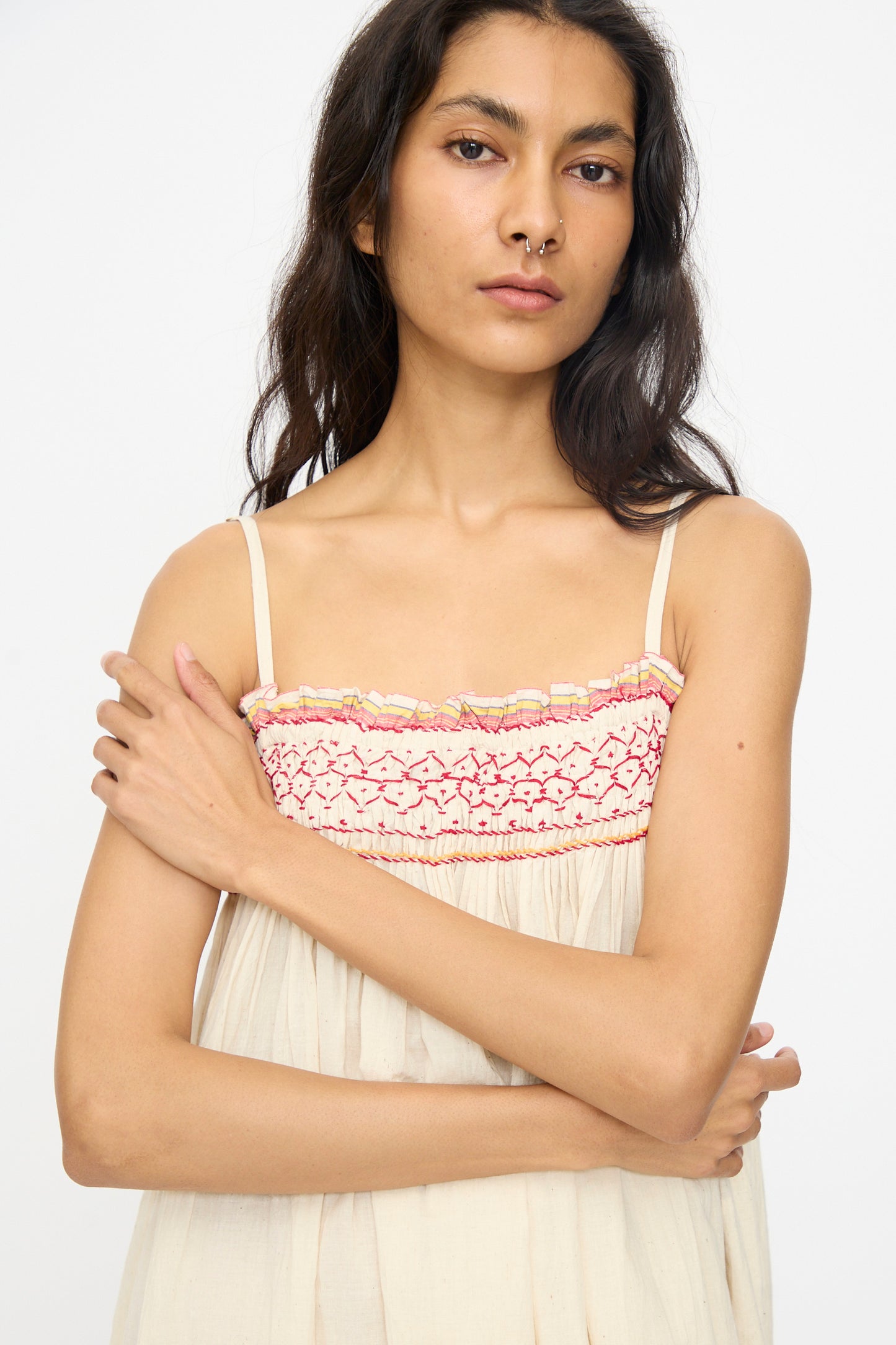 A woman with long dark hair models the Injiri Woven Cotton Slip Dress in White with Red Stitch, standing with one arm crossed and looking at the camera against a plain background.