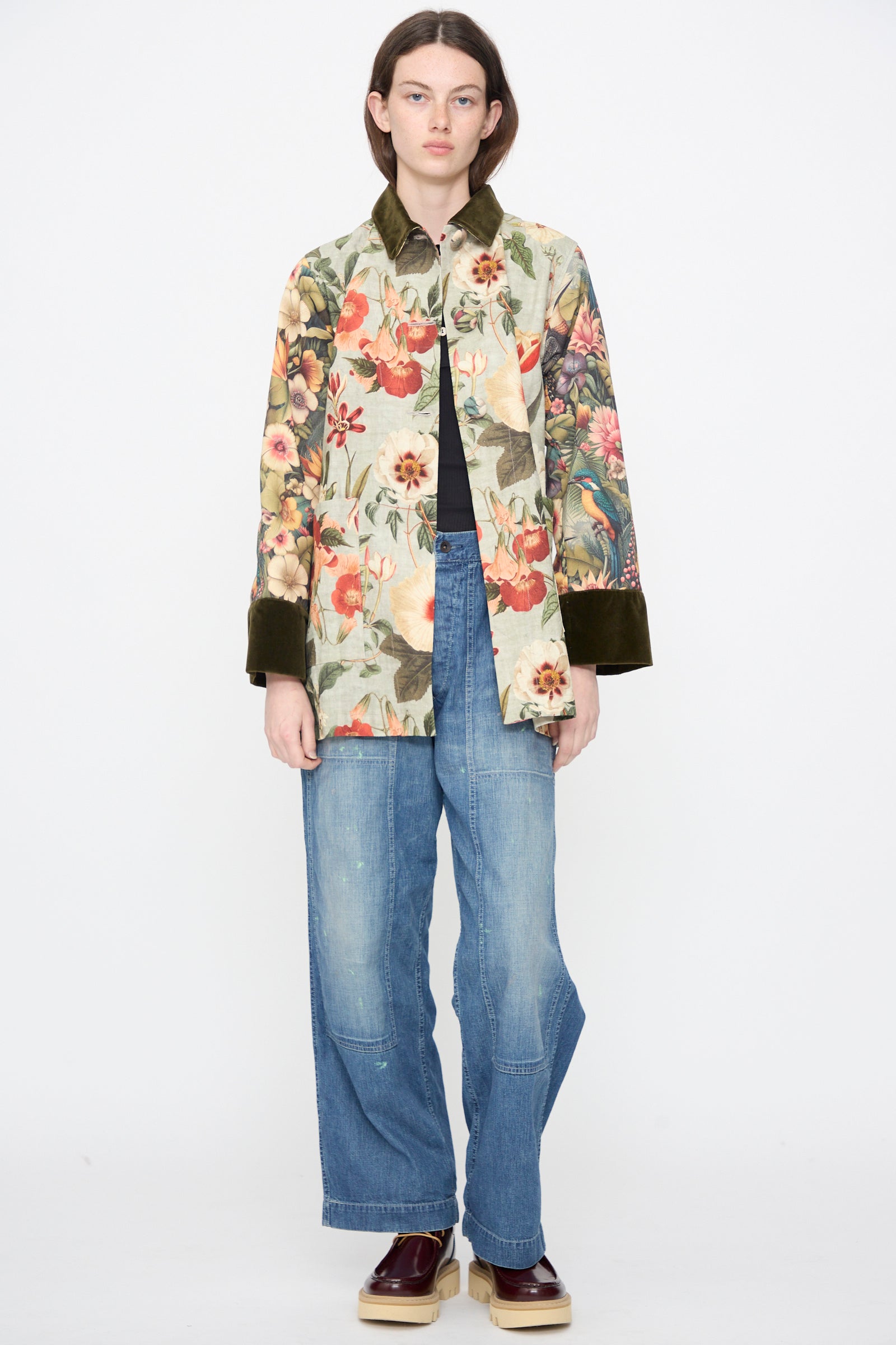 A person stands against a white background wearing the Intensity Cotton Coat with Velvet in Green Floral, wide-leg blue jeans, a black top, and brown platform shoes.