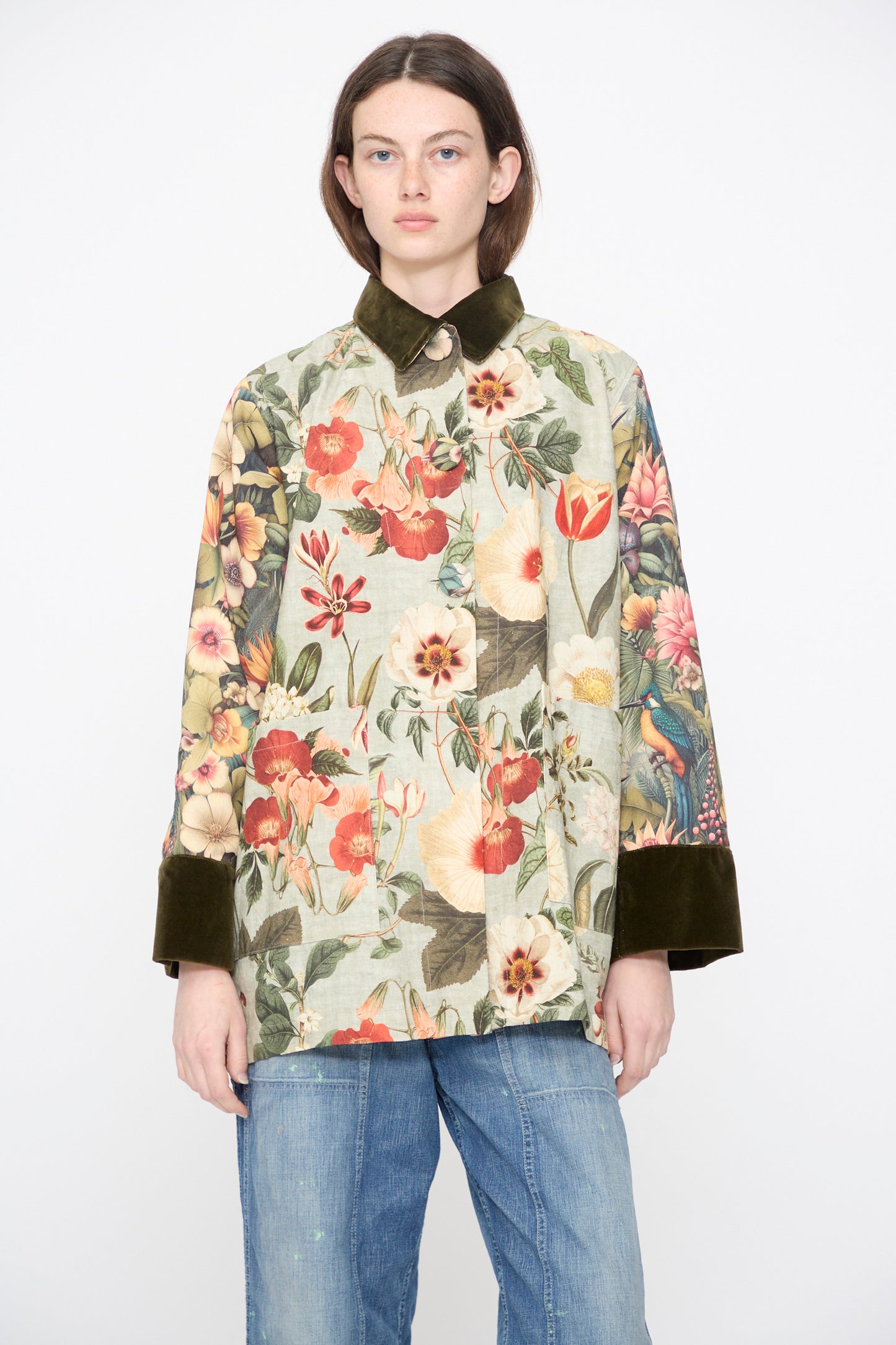 A model wears the Intensity Cotton Coat with Velvet in Green Floral, featuring a velvet collar and dark cuffs, paired with blue jeans, against a plain white background.