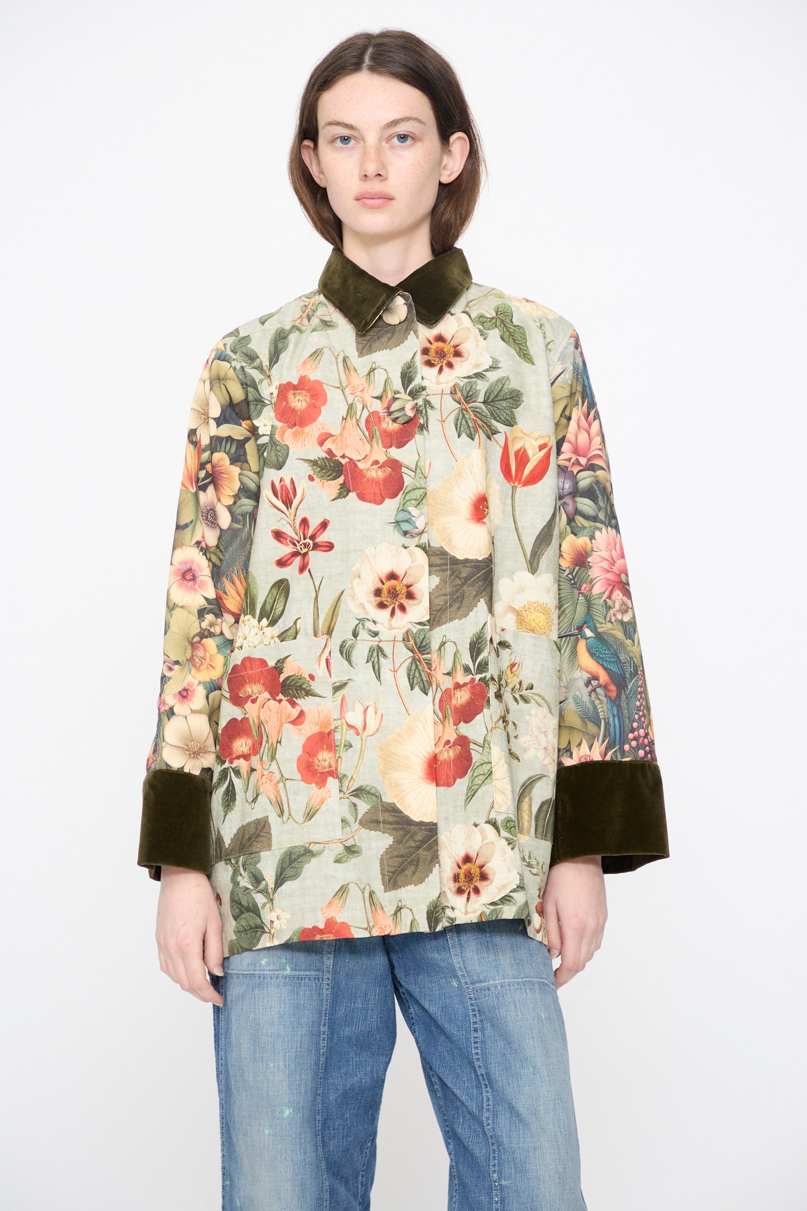 A model wears the Intensity Cotton Coat with Velvet in Green Floral, featuring a velvet collar and dark cuffs, paired with blue jeans, against a plain white background.
