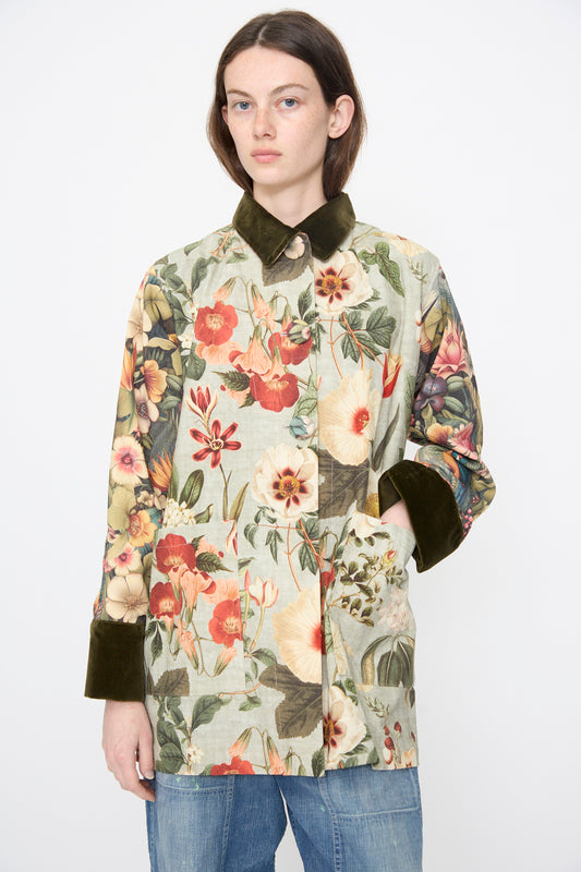 A person wears the Intensity Cotton Coat with Velvet in Green Floral, featuring a dark velvet collar and cuffs, styled with blue jeans against a plain white background.