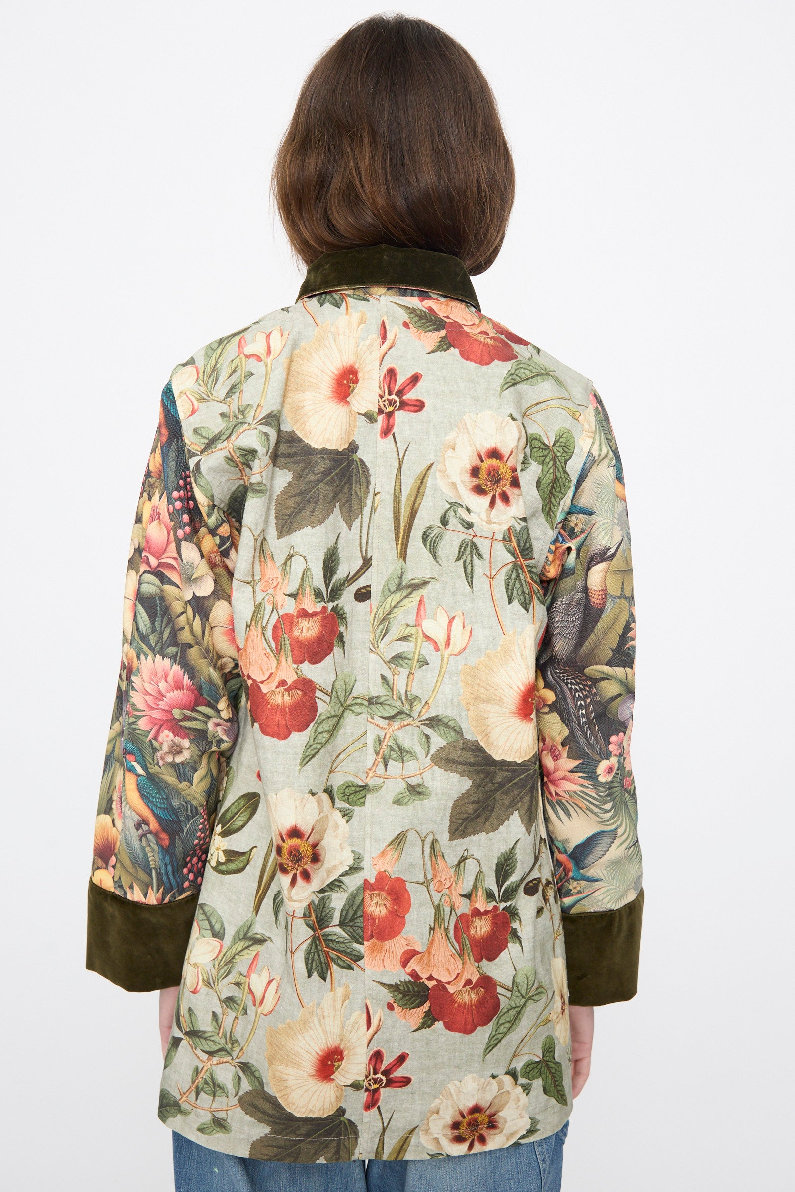 A person facing away wears the Intensity Cotton Coat with Velvet in Green Floral, featuring long sleeves, a colorful floral and bird print, and a dark velvet collar.