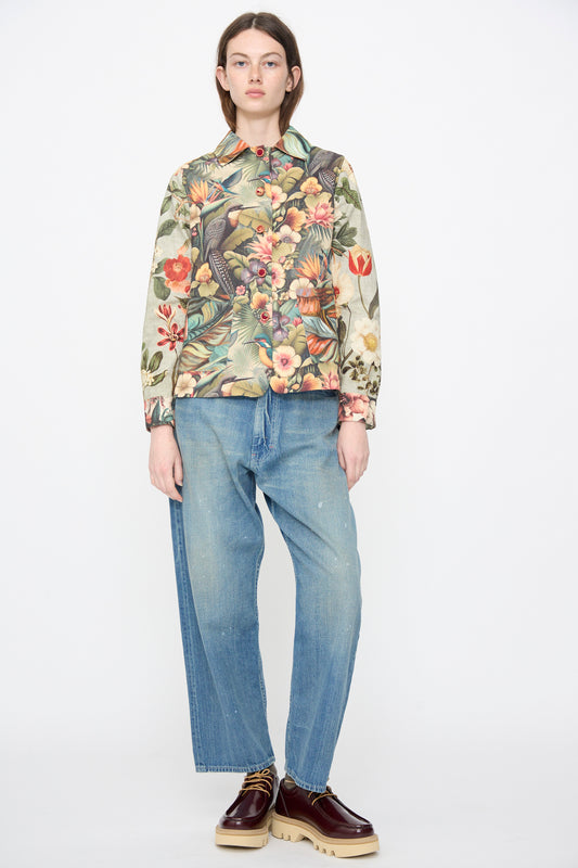A person stands facing forward against a plain white background, wearing Intensity’s Cotton Shirt Jacket in Green Floral, loose blue jeans, and brown platform shoes.