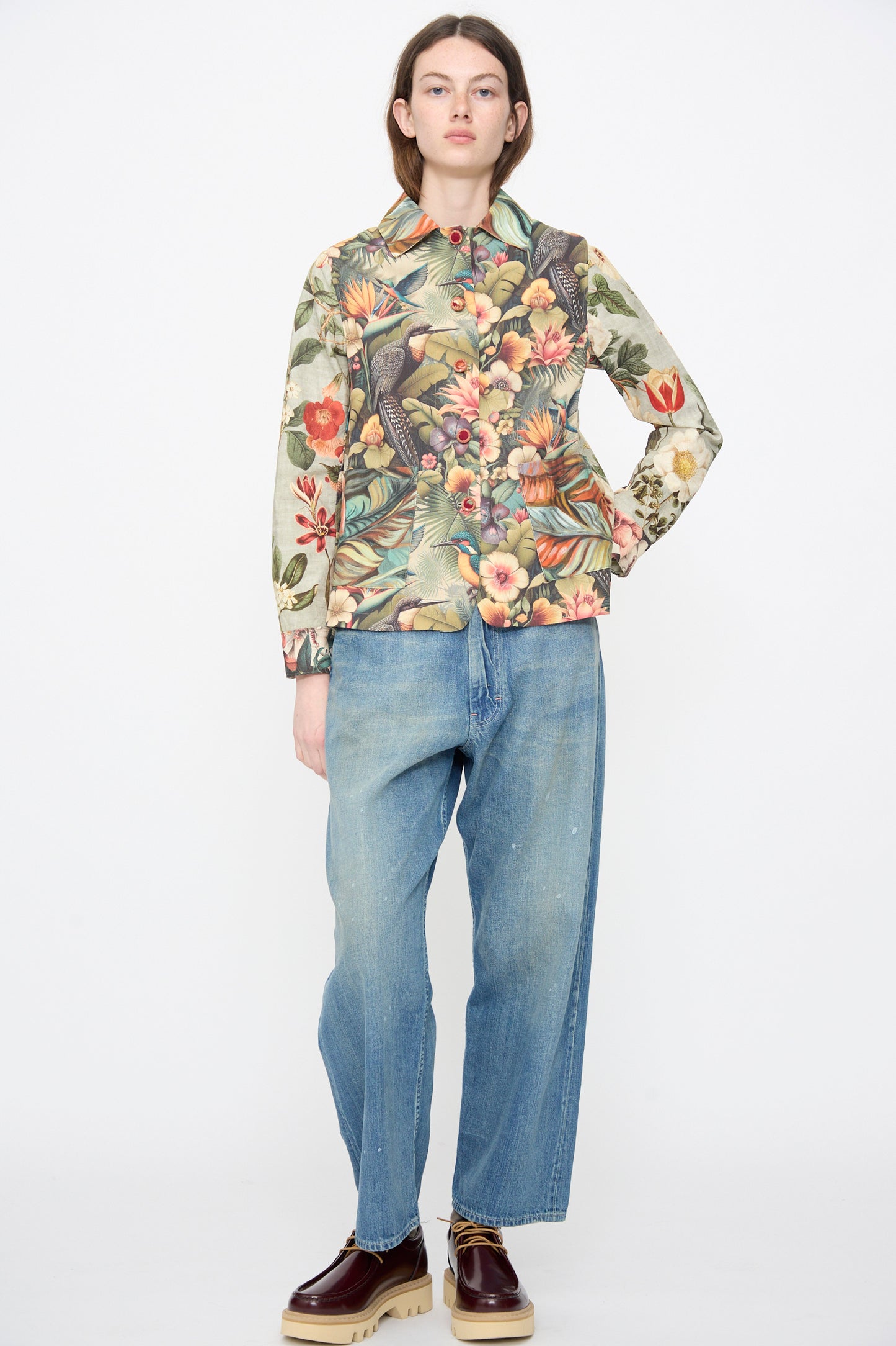 A person stands against a white background, wearing the Intensity Cotton Shirt Jacket in Green Floral, loose blue jeans, and brown platform shoes.