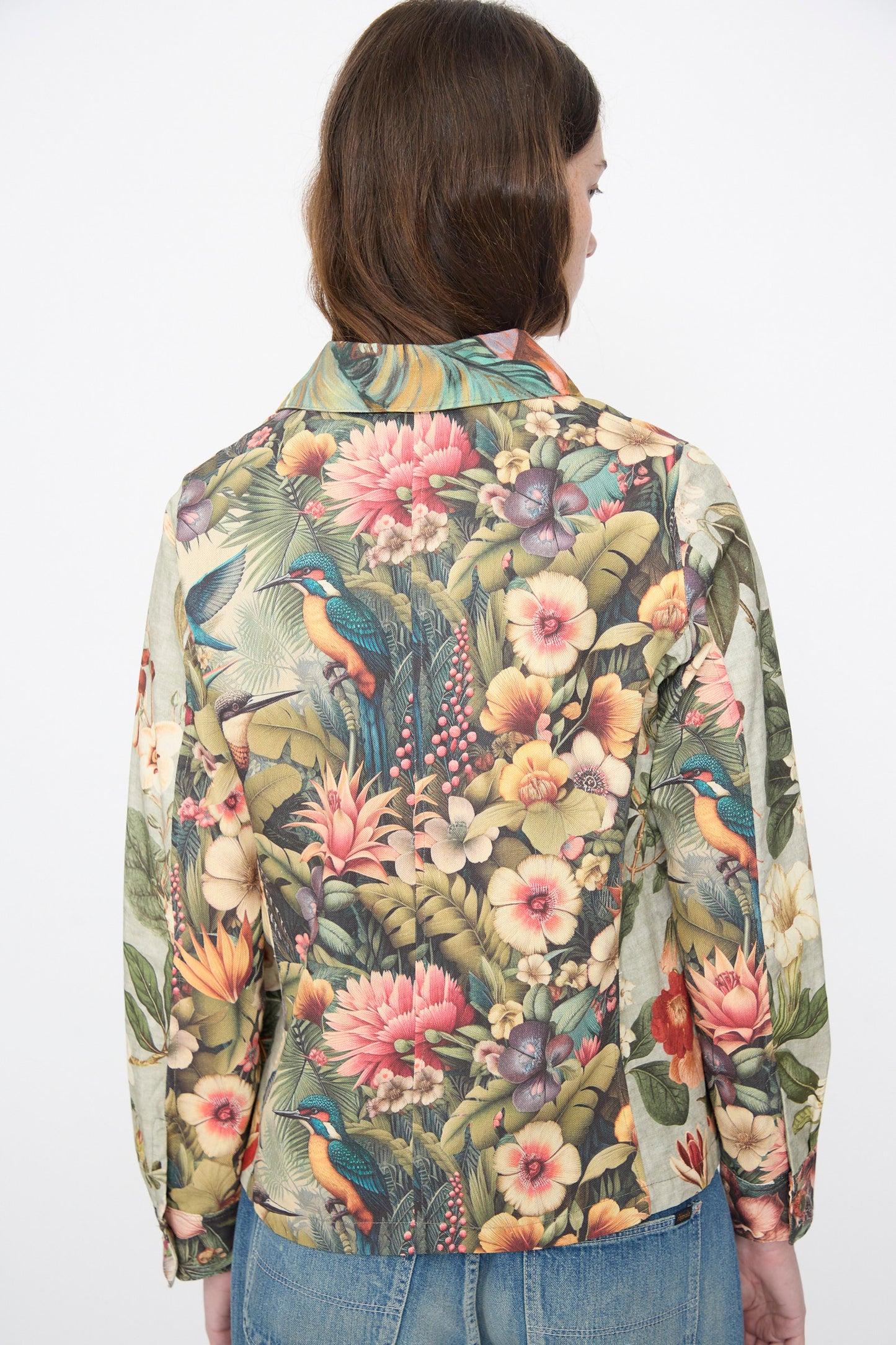Someone is seen from behind wearing the Intensity Cotton Shirt Jacket in Green Floral—a cotton piece featuring a tropical bird and floral print—paired with blue jeans, set against a plain white background.