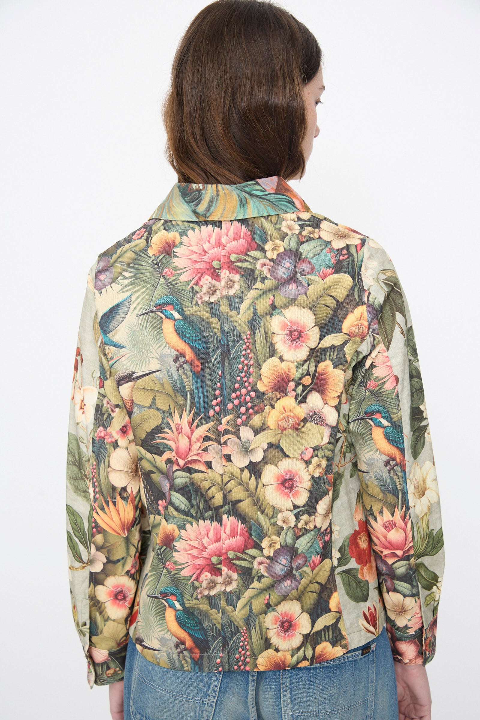Someone is seen from behind wearing the Intensity Cotton Shirt Jacket in Green Floral—a cotton piece featuring a tropical bird and floral print—paired with blue jeans, set against a plain white background.
