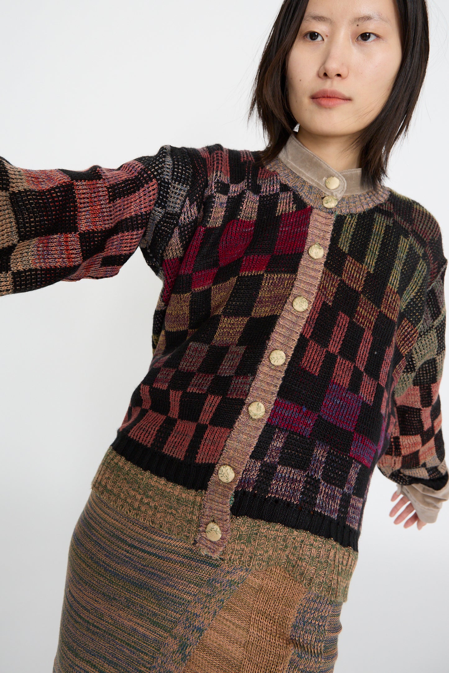 Person dressed in the Intensity Optic Cardigan in Multi I, featuring a multicolored checkered design with velvet trim and buttons, paired with a matching skirt, against a white backdrop. 