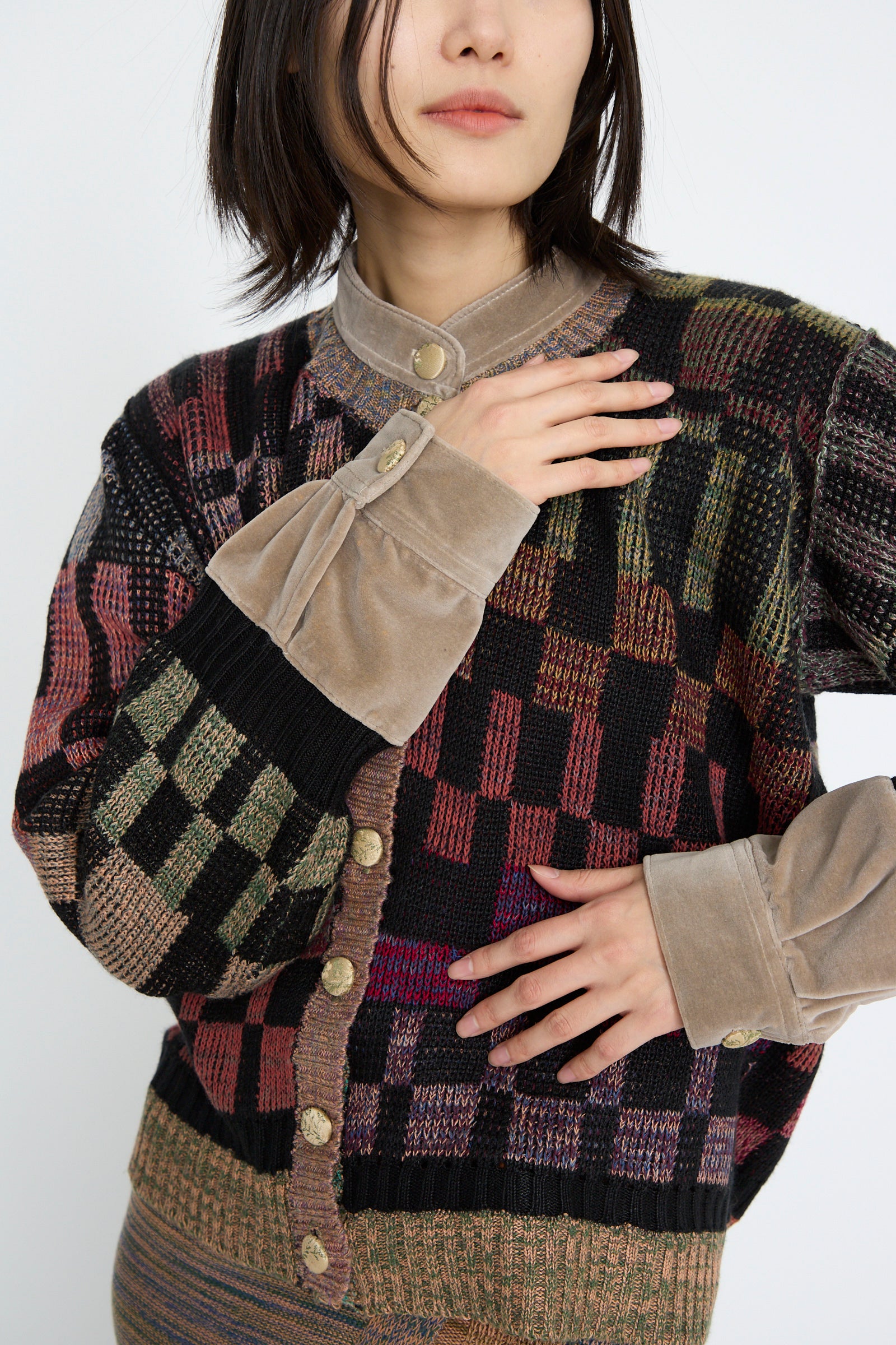 A person wearing the Intensity Optic Cardigan in Multi I, showcasing its graphic knit design, large buttons, and beige velvet trim. They pose with one hand on the collar and the other on their chest against a plain background.