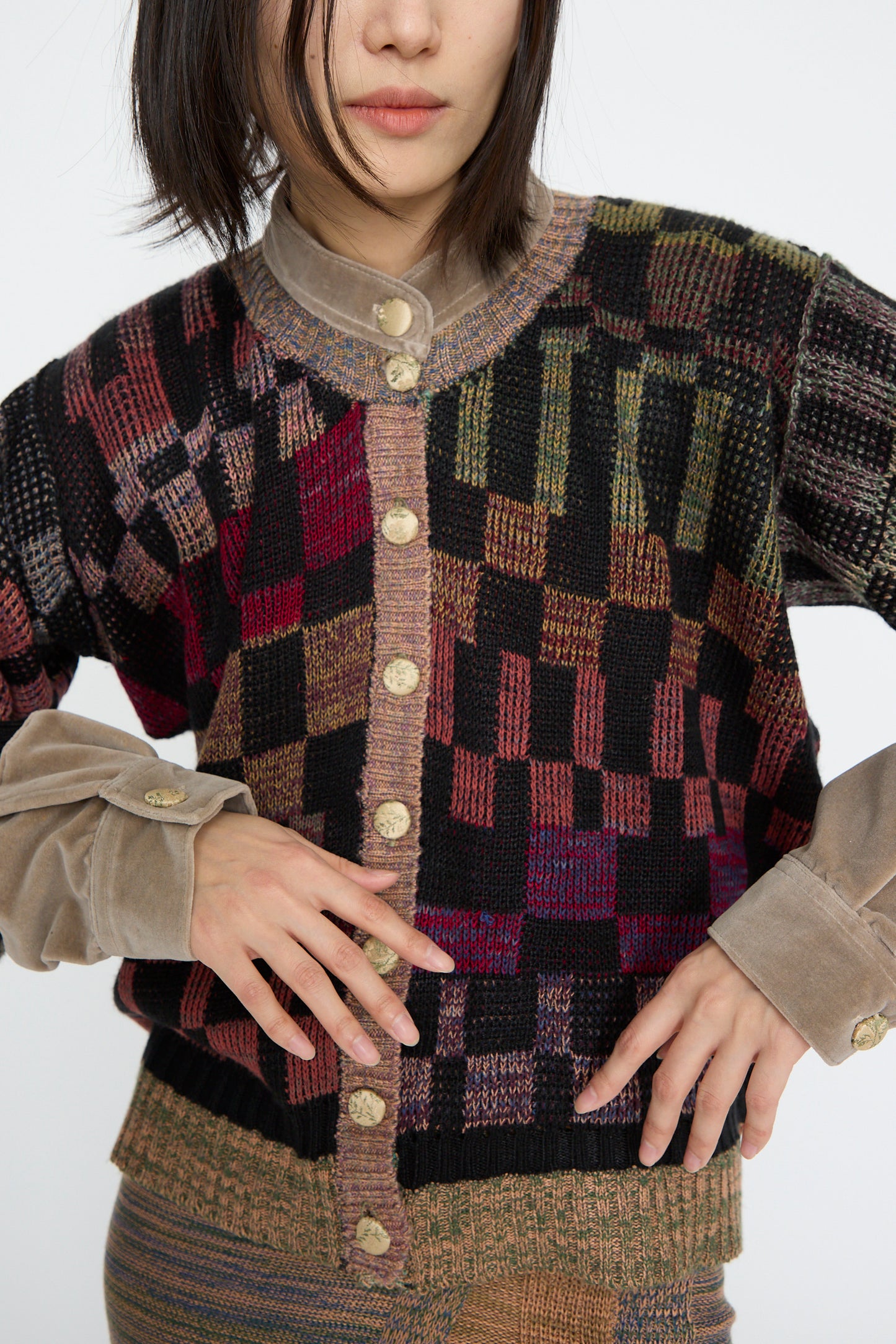 A person wearing the Intensity Optic Cardigan in Multi I, a vibrant patterned cardigan with a button-down front over a beige collared shirt, poses with hands on their waist.