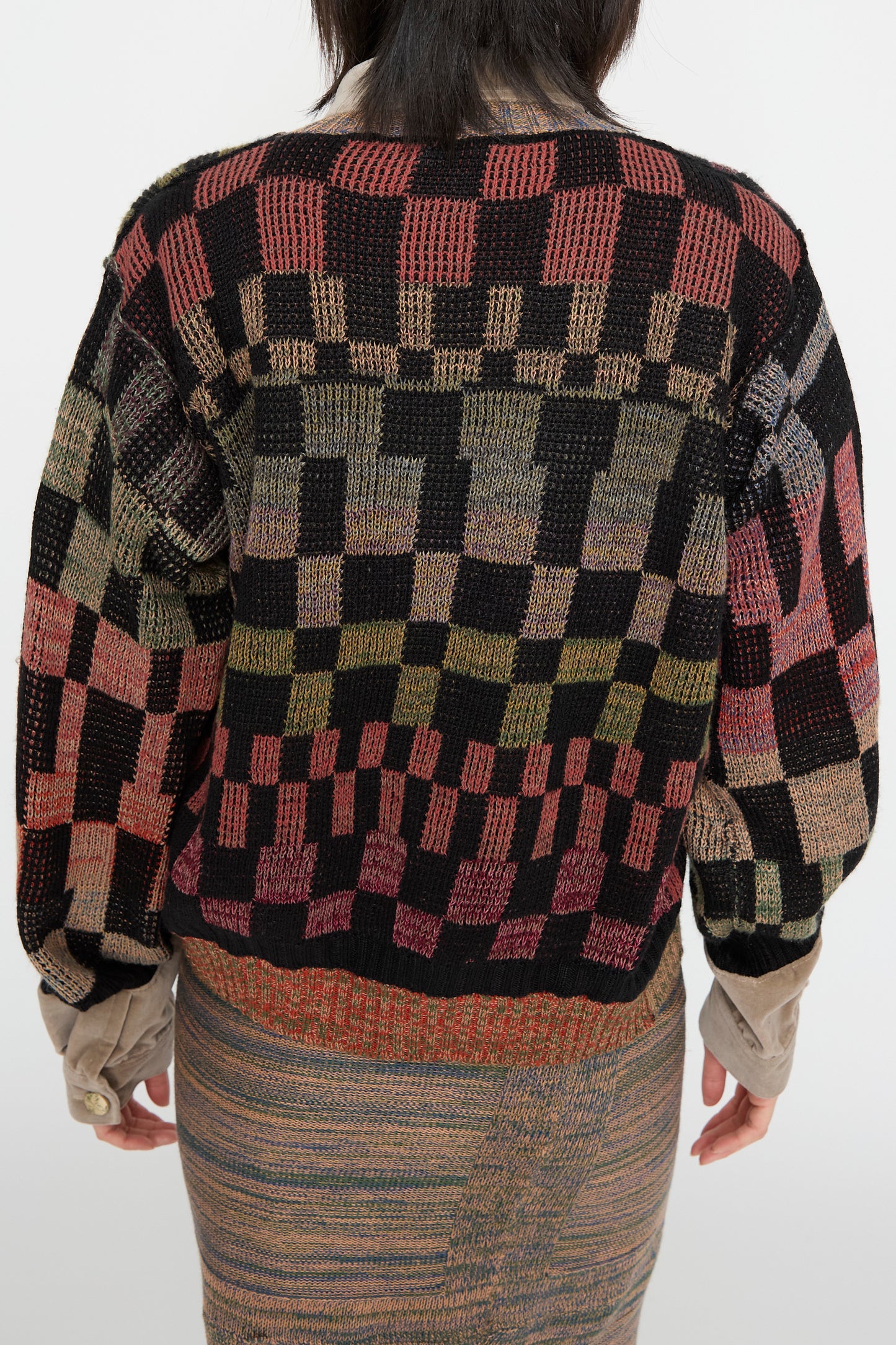 A person is wearing the Optic Cardigan in Multi I by Intensity, featuring a colorful checkered design with long sleeves and a patterned skirt with subtle velvet trim, while facing away.