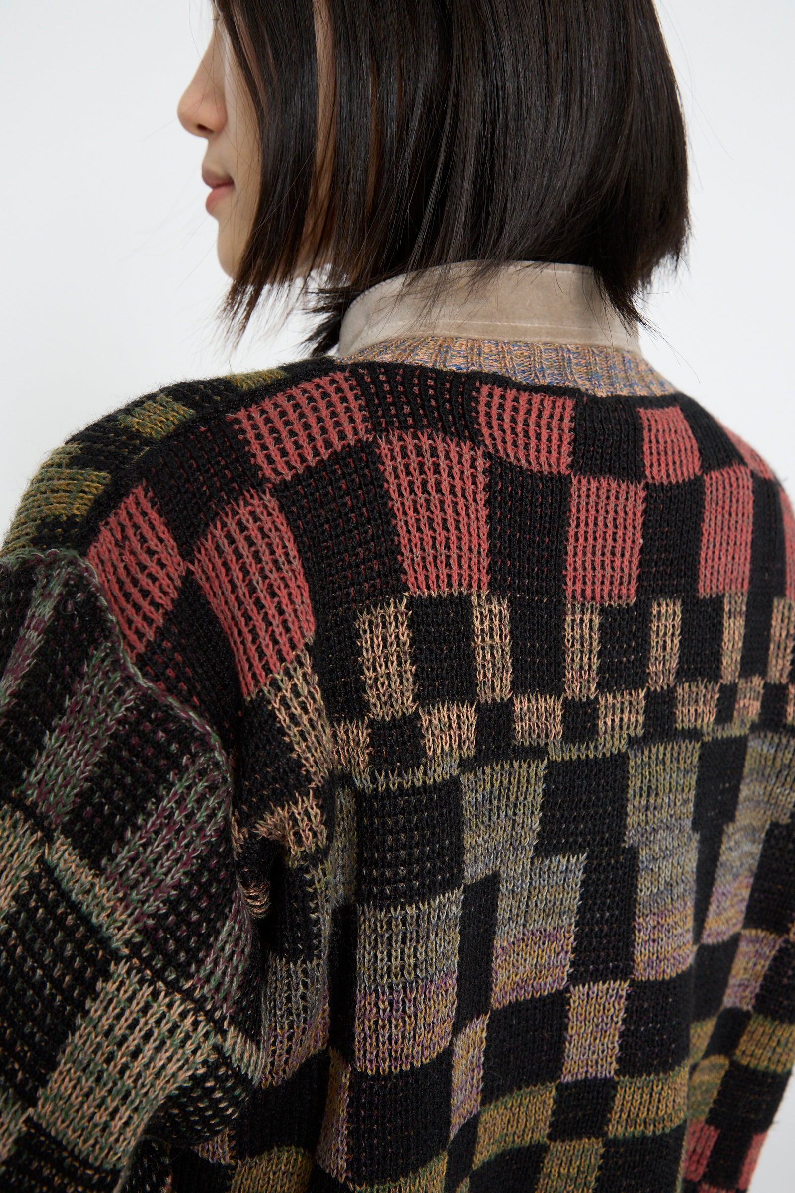 A person wearing the Intensity Optic Cardigan in Multi I, featuring a multi-colored checkered knit pattern and velvet trim, layered over a collared shirt, viewed from the side and back.