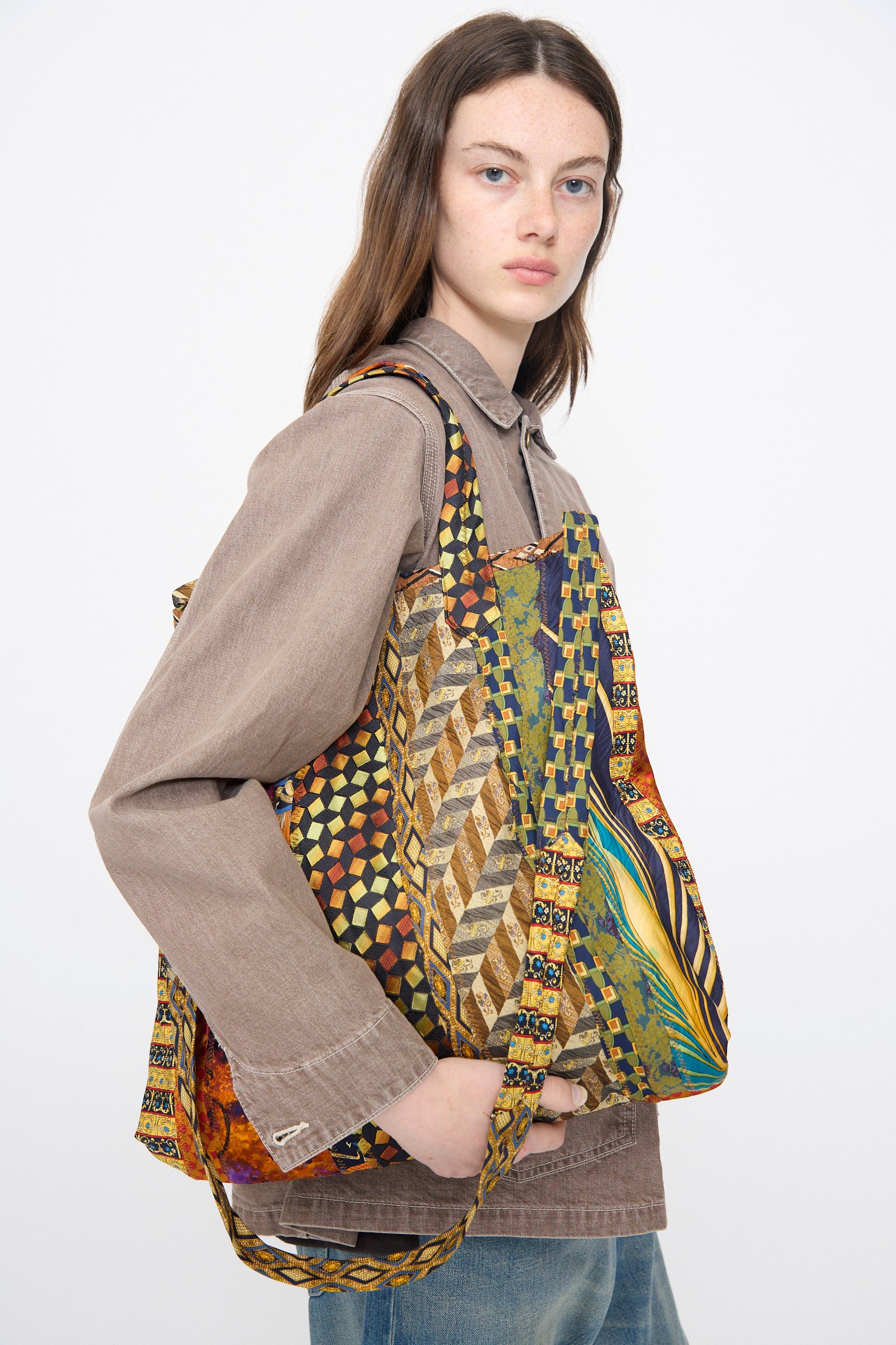 A woman with long brown hair wears a taupe button-up and blue jeans, carrying the Intensity Ties Bag in Orange Multi—this unique, colorful tote adds a bold, stylish touch to her everyday look.