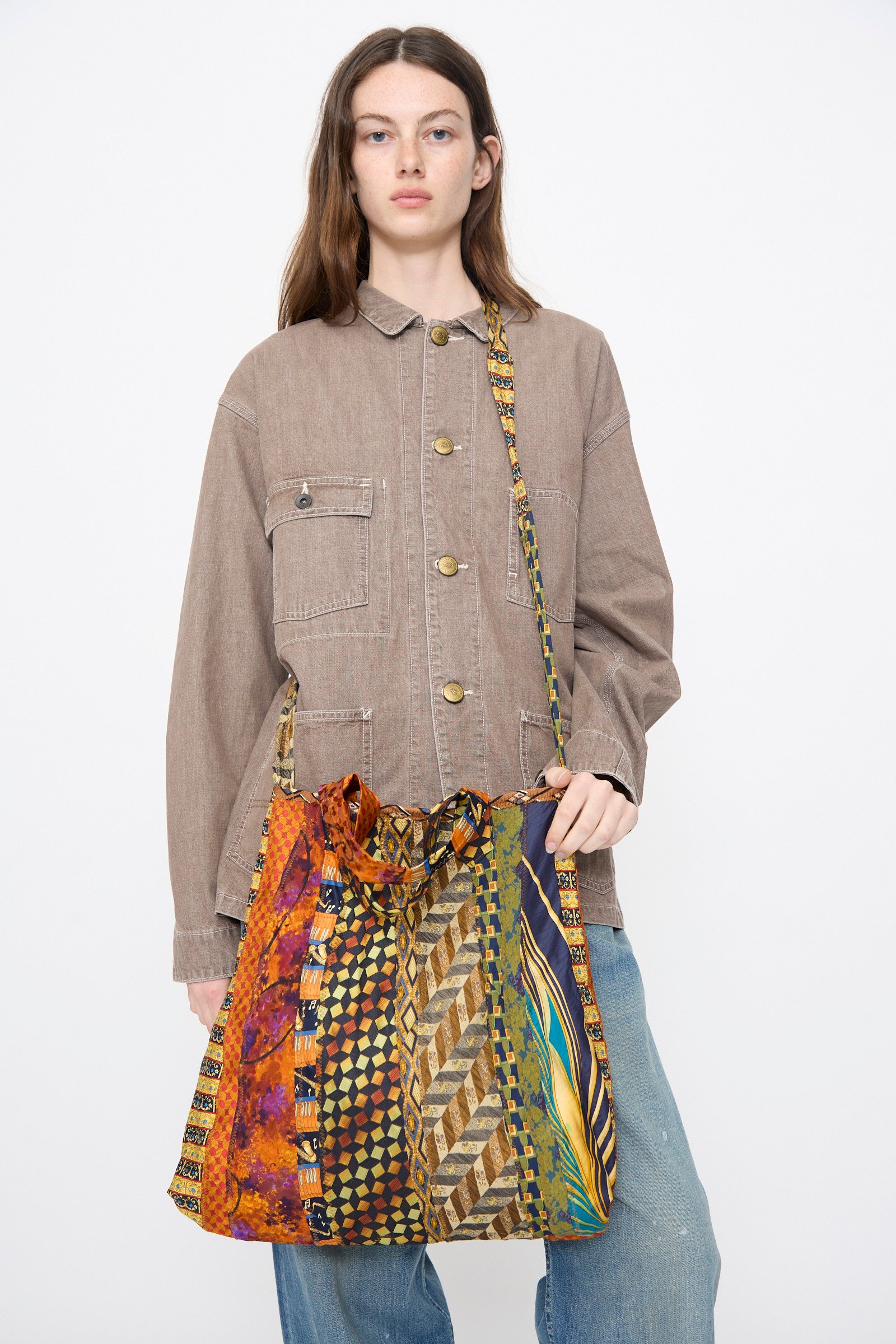 A person wearing a tan button-up jacket and blue jeans carries the Intensity Ties Bag in Orange Multi—a large, multicolored patchwork shoulder bag with a patterned strap.