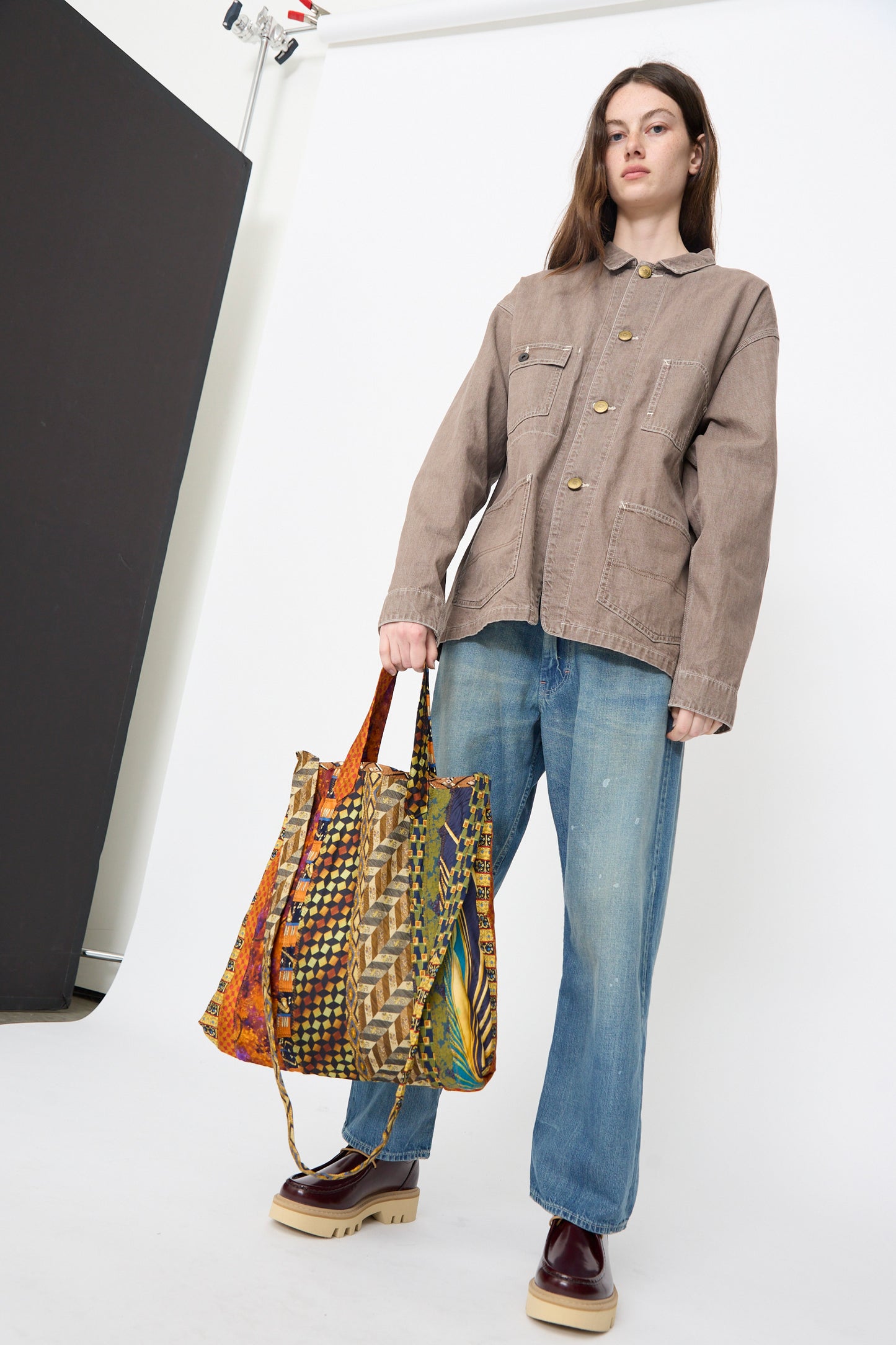 A person stands in a studio wearing a brown jacket, blue jeans, and platform shoes, holding the Intensity Ties Bag in Orange Multi.