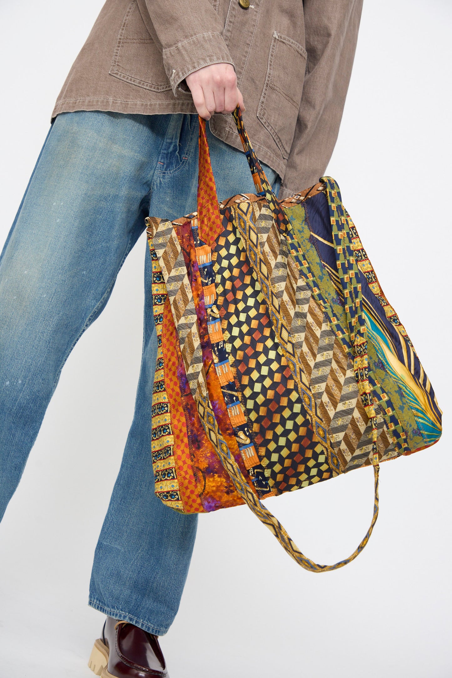 A person in blue jeans and a brown jacket carries the Intensity Ties Bag in Orange Multi—a large tote crafted from patterned silk ties featuring mixed geometric and abstract designs.