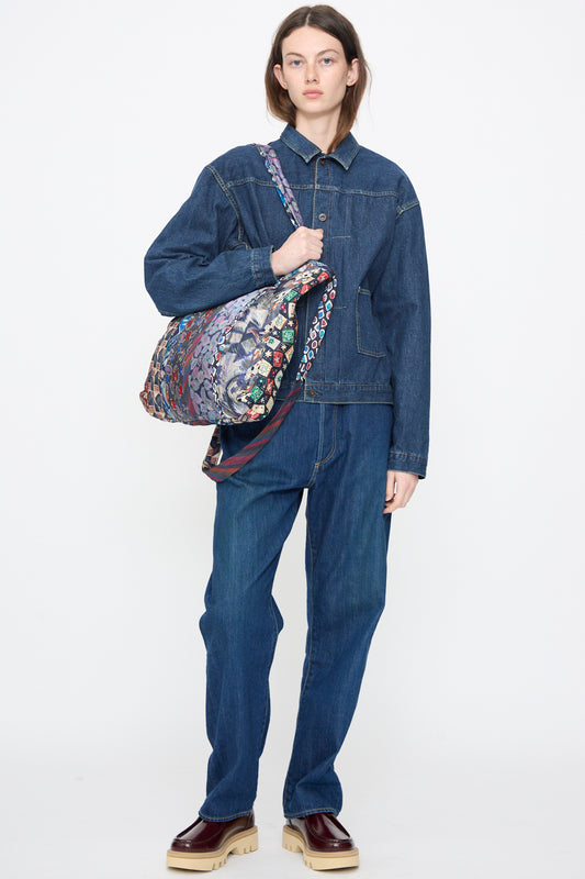 A person wearing a denim jacket and jeans holds the Intensity Ties Bag in Purple Multi on their shoulder, standing against a plain white background.