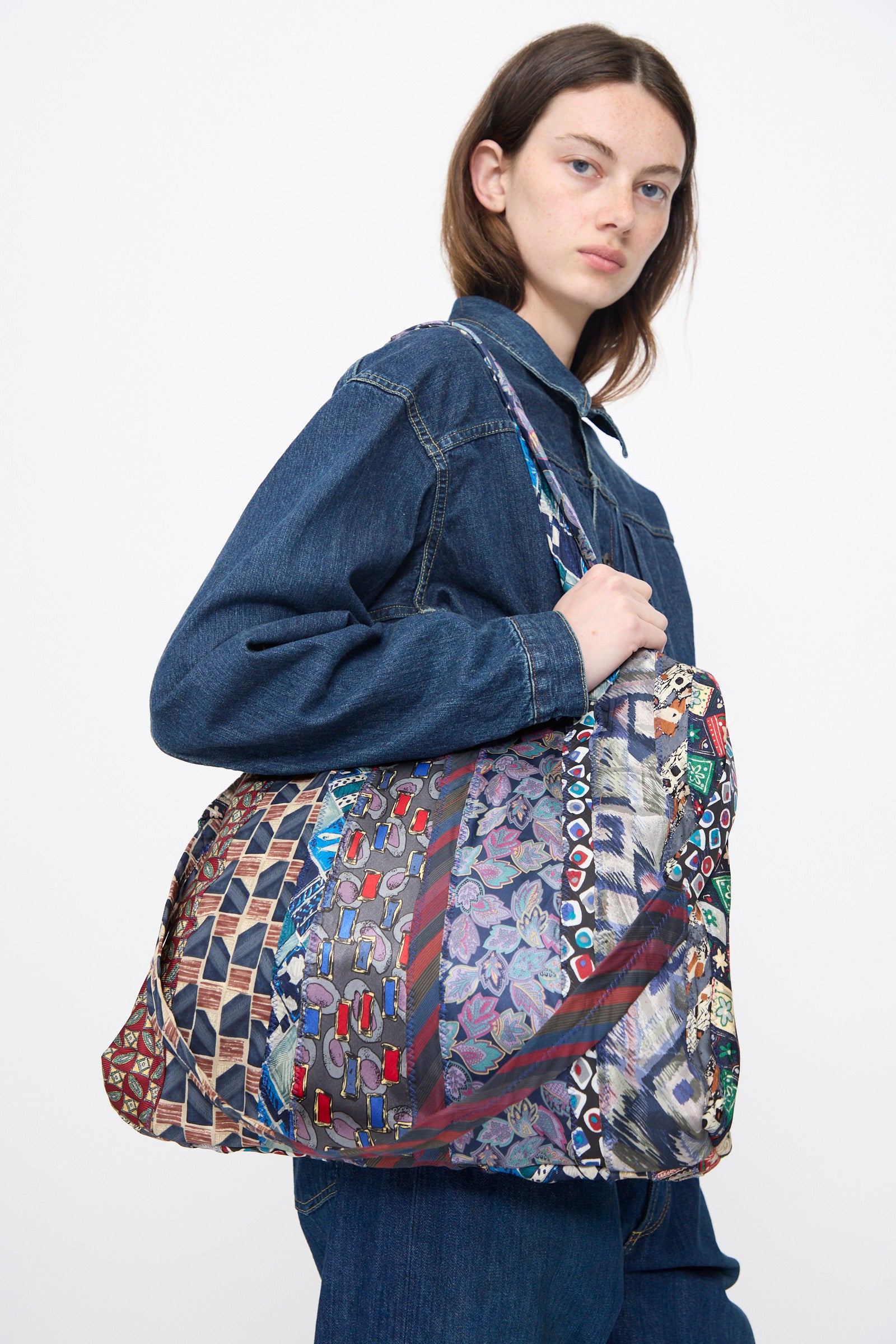 A woman in a denim jacket and jeans carries the Intensity Ties Bag in Purple Multi—a large, vintage-style silk tote with a multicolored pattern—over her shoulder against a plain background.