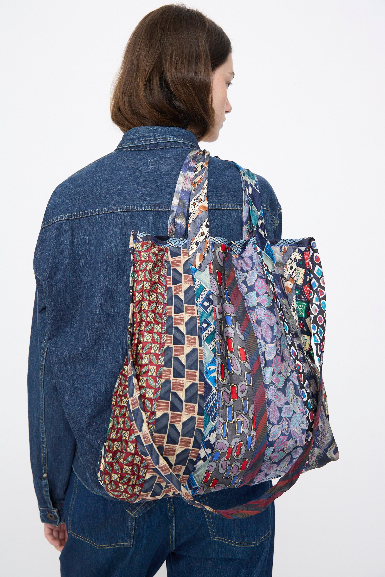 A person wearing a denim jacket and jeans carries the Intensity Ties Bag in Purple Multi, a large vintage silk tote with multi-patterned fabrics, over their shoulder, facing away from the camera.