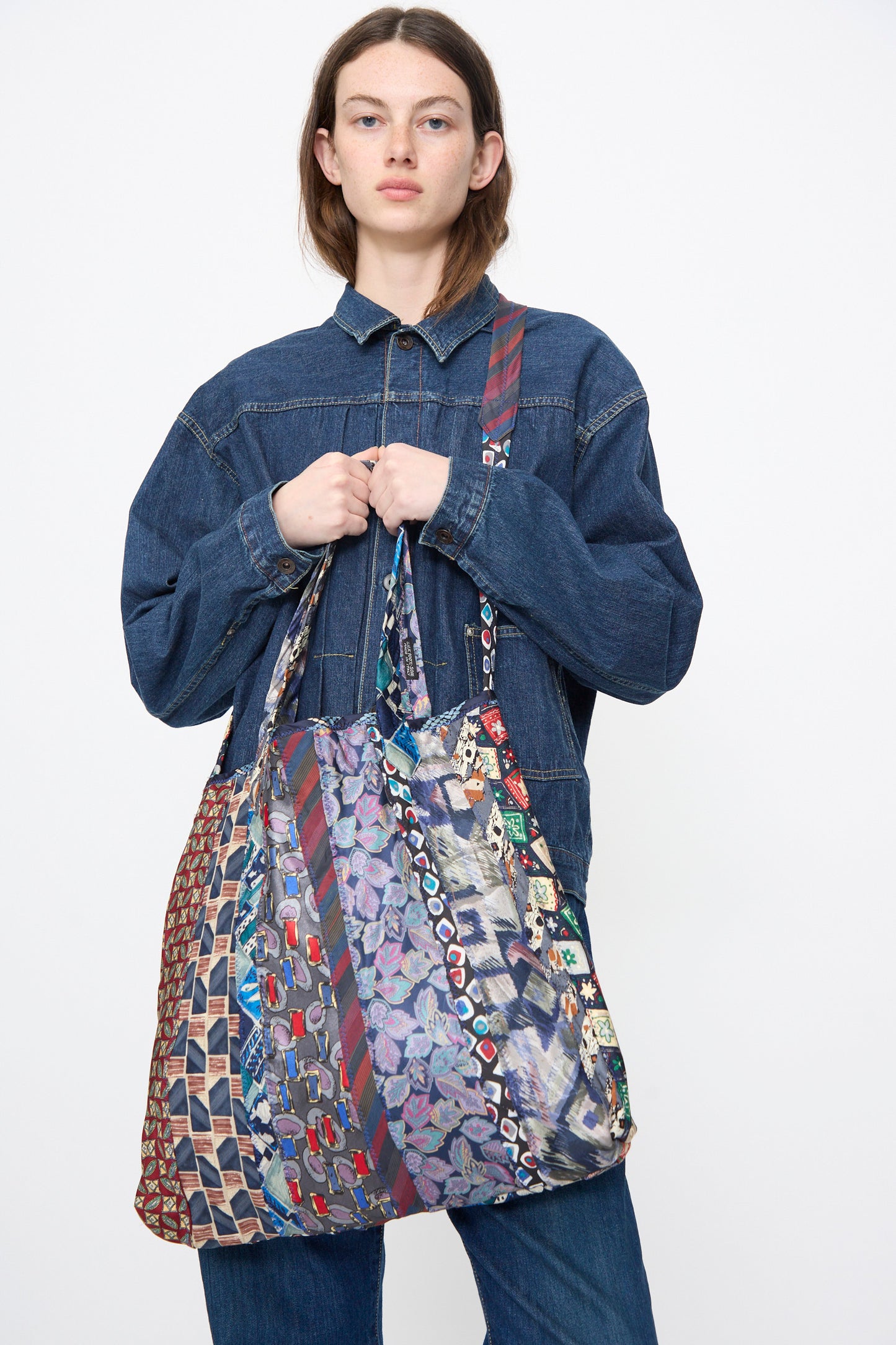A person in a denim jacket and jeans holds the Intensity Ties Bag in Purple Multi, a large vintage silk tote with diagonal stripes and colorful designs.