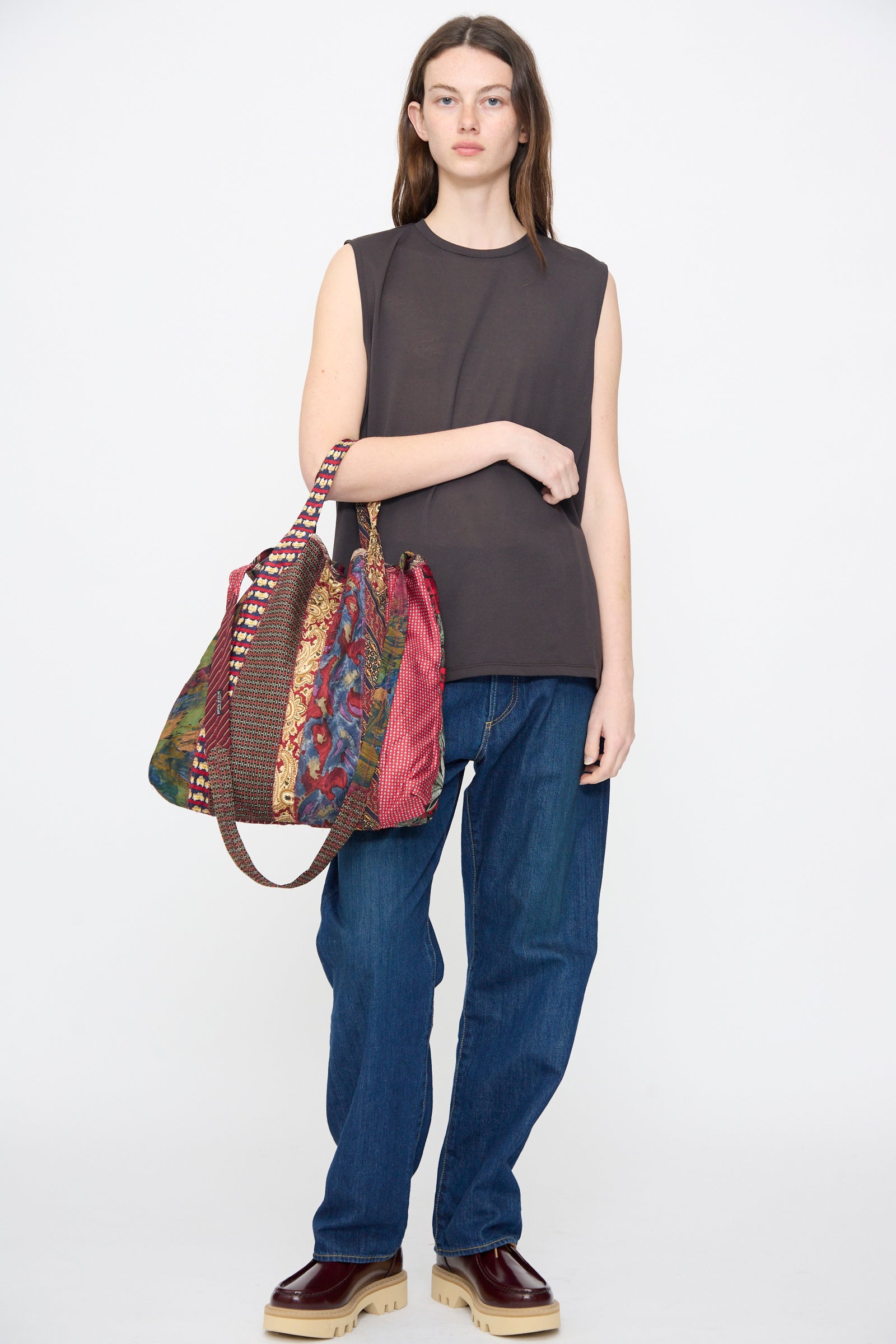 A person in a sleeveless black shirt, blue jeans, and maroon shoes holds the Intensity Ties Bag in Red Multi—a distinctive vintage-style tote—against a plain white background.