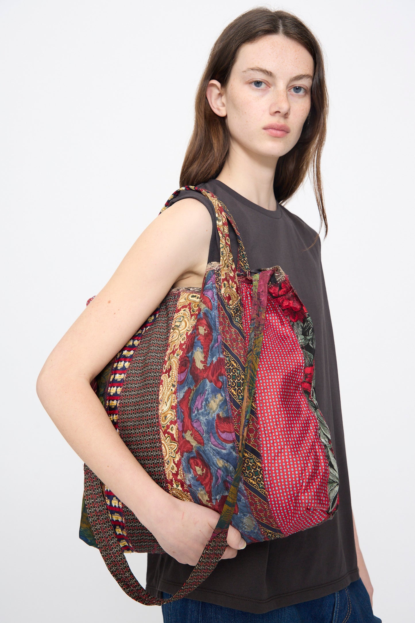 A person with long brown hair wears a sleeveless dark top and carries the Intensity Ties Bag in Red Multi, a unique vintage silk tote with multiple patterns, over their shoulder.
